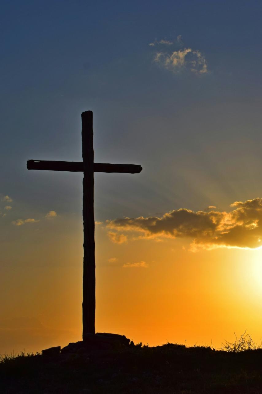 Auf einem Berg steht ein großes Kreuz, dahinter geht die Sonne unter