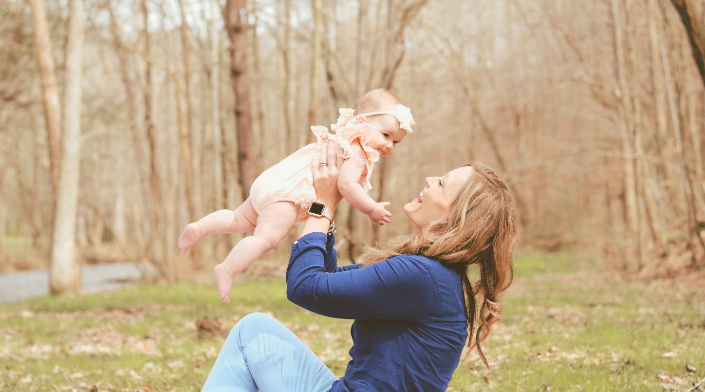 Eine Frau sitzt auf einer Wiese und hält lächelnd ein Baby hoch in die Luft
