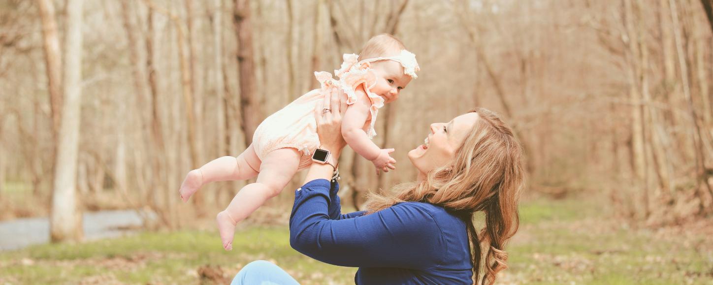 Eine Frau sitzt auf einer Wiese und hält lächelnd ein Baby hoch in die Luft
