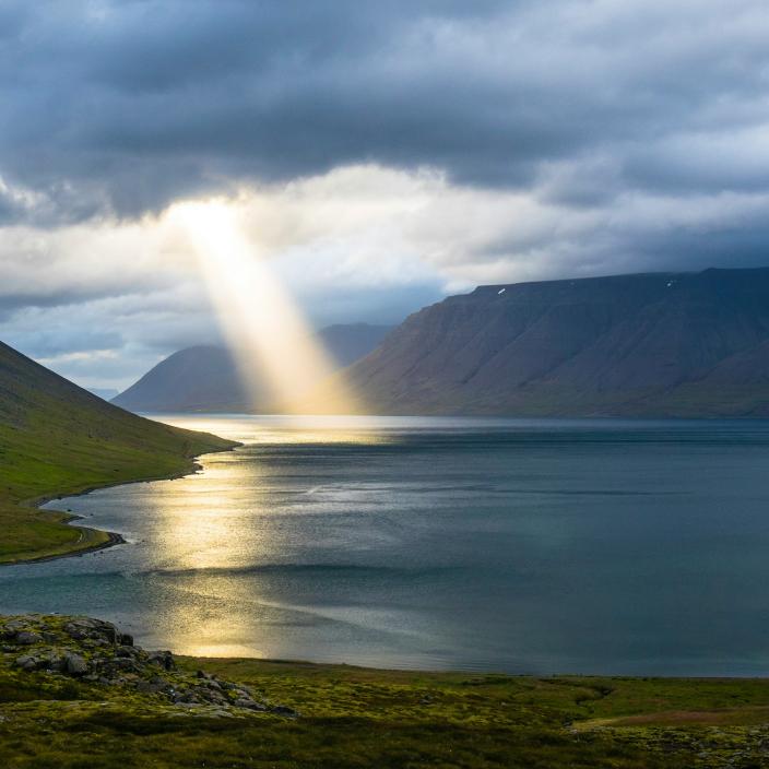 Ein Lichtstrahl der Sonne bricht durch Wolken und trifft ein Gewässer. Dahinter sind Berge zu sehen