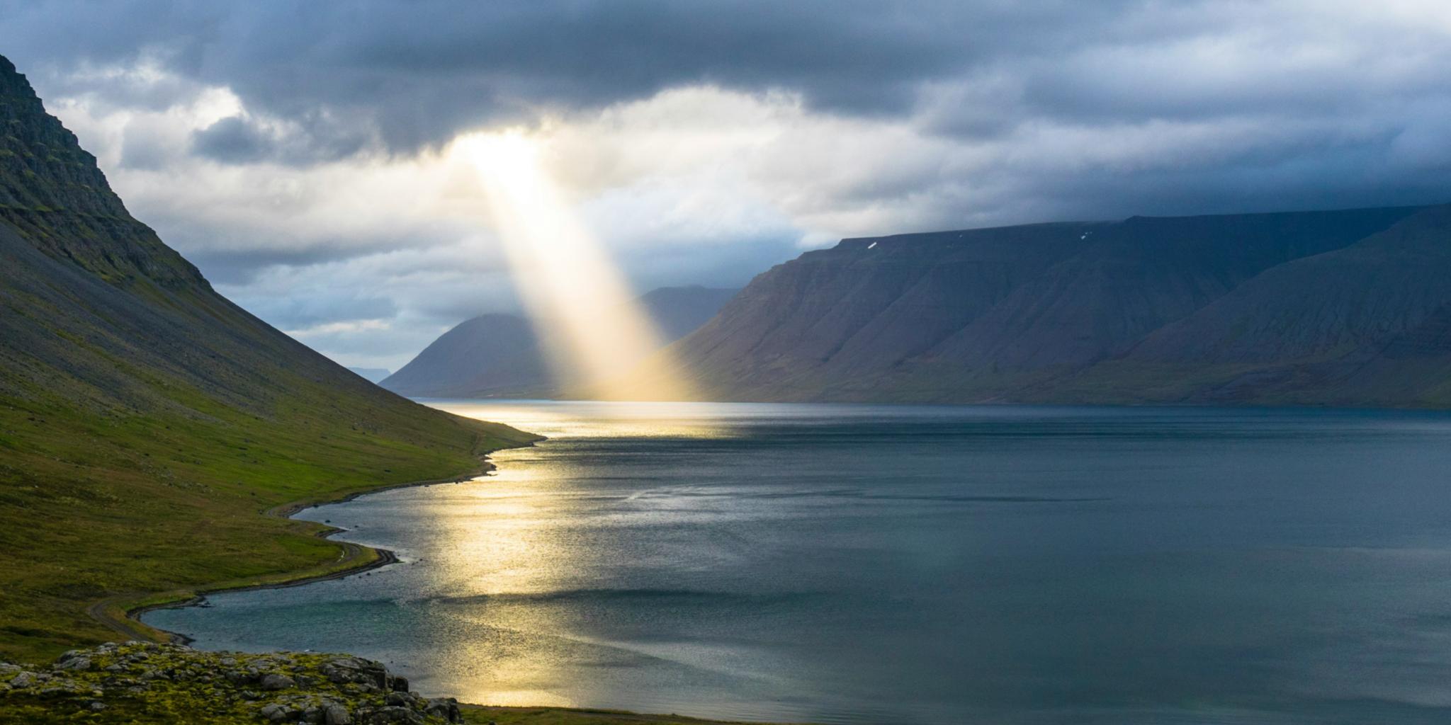 Ein Lichtstrahl der Sonne bricht durch Wolken und trifft ein Gewässer. Dahinter sind Berge zu sehen
