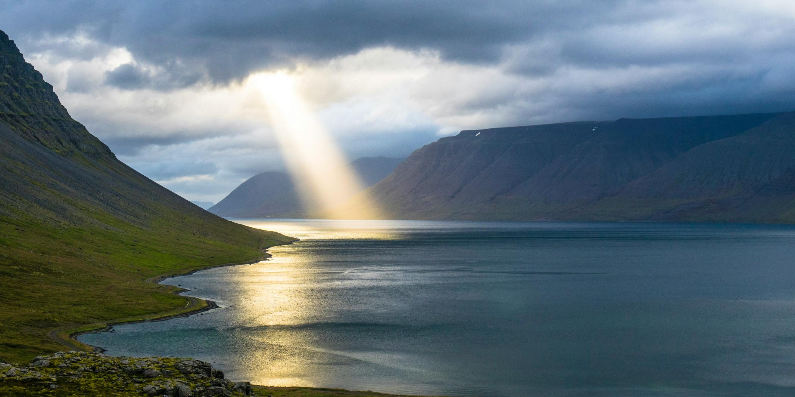 Ein Lichtstrahl der Sonne bricht durch Wolken und trifft ein Gewässer. Dahinter sind Berge zu sehen