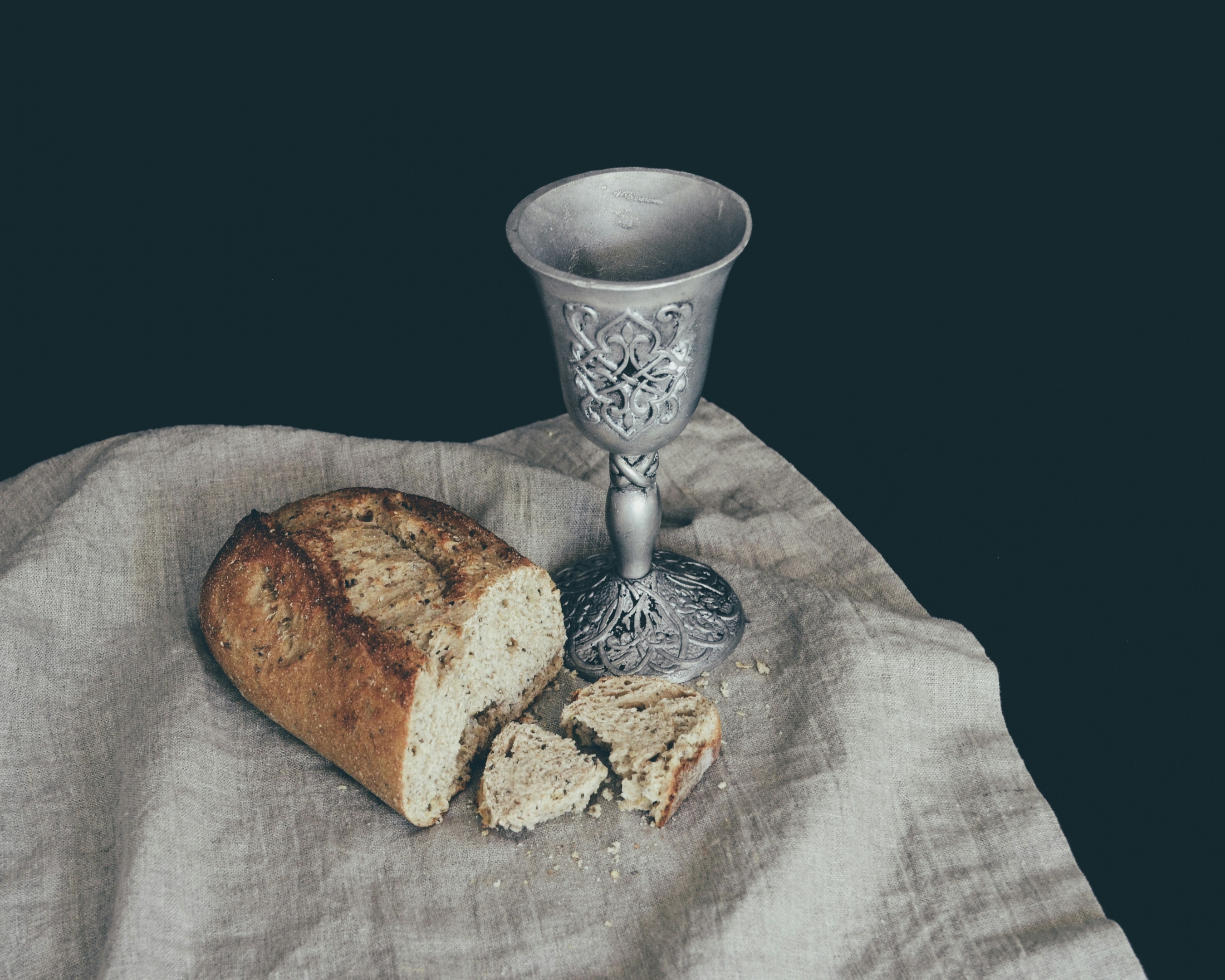 Auf einem Leinentuch liegt ein angeschnittener Laib Brot. Daneben steht ein Kelch mit Verzierungen in mattem Silber