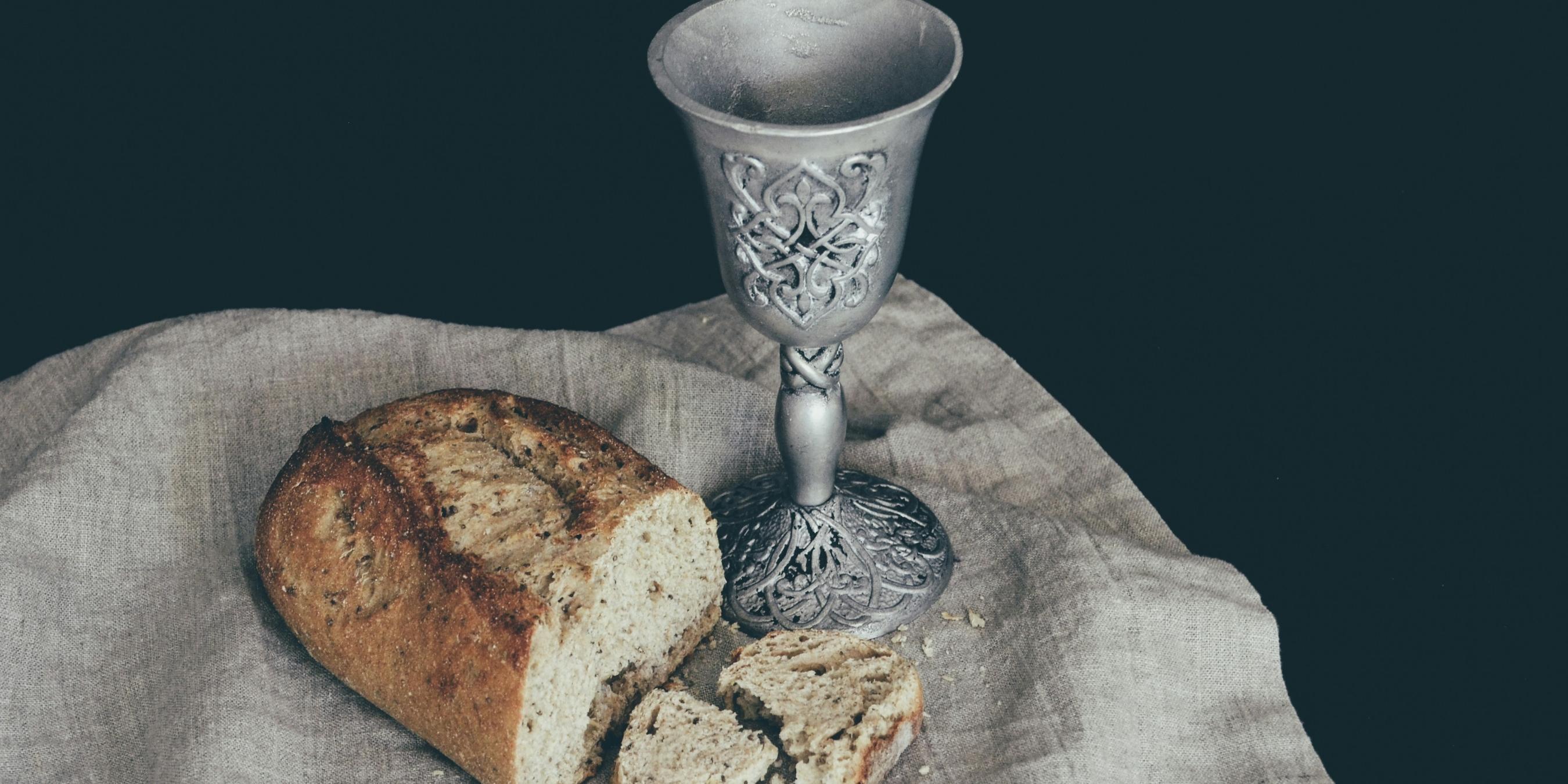 Auf einem Leinentuch liegt ein angeschnittener Laib Brot. Daneben steht ein Kelch mit Verzierungen in mattem Silber