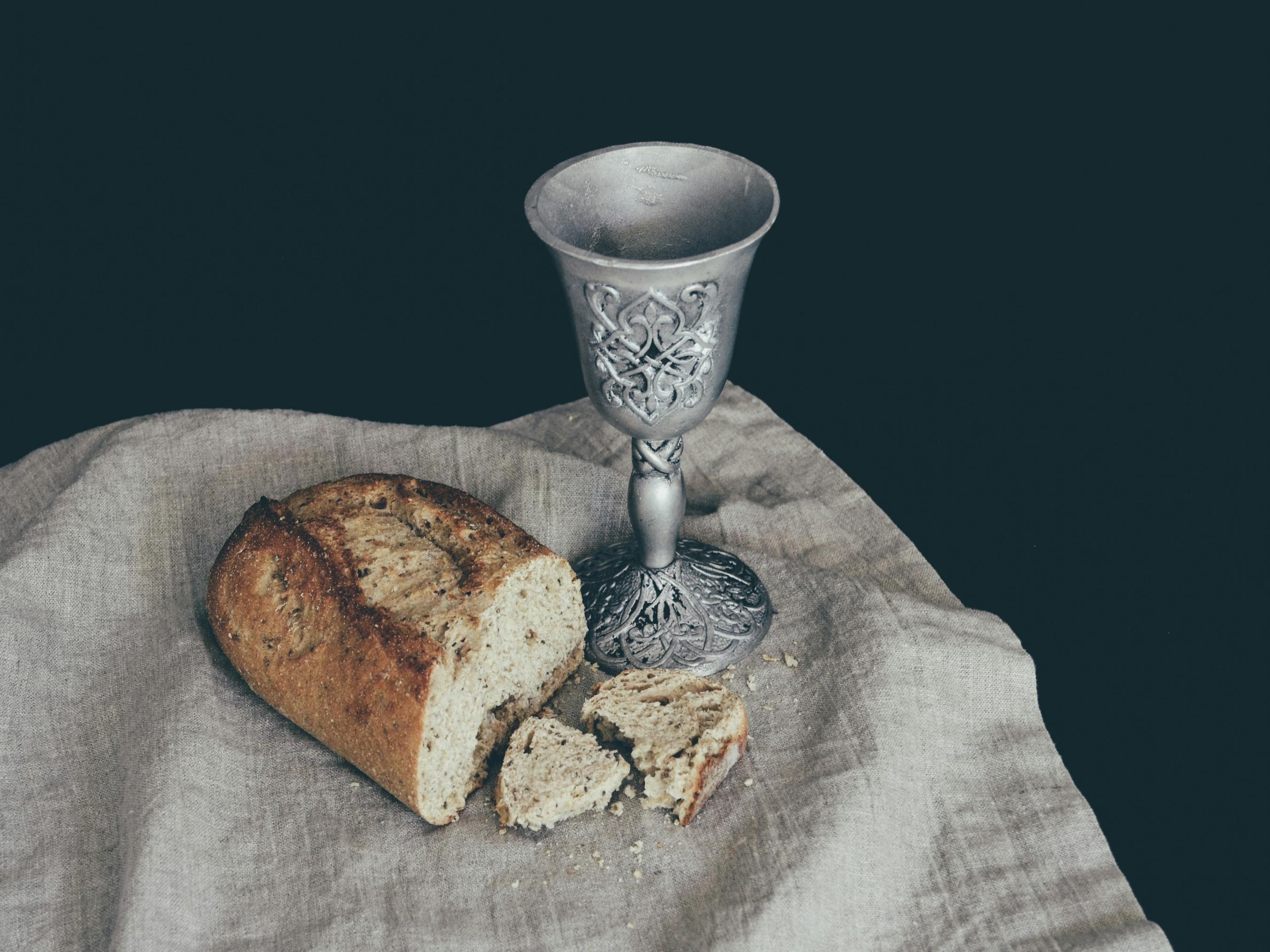 Auf einem Leinentuch liegt ein angeschnittener Laib Brot. Daneben steht ein Kelch mit Verzierungen in mattem Silber