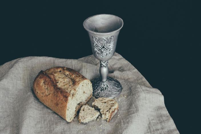 Auf einem Leinentuch liegt ein angeschnittener Laib Brot. Daneben steht ein Kelch mit Verzierungen in mattem Silber