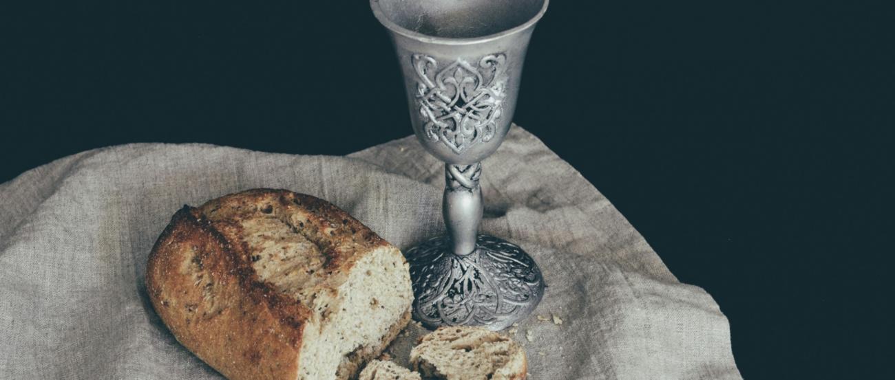 Auf einem Leinentuch liegt ein angeschnittener Laib Brot. Daneben steht ein Kelch mit Verzierungen in mattem Silber