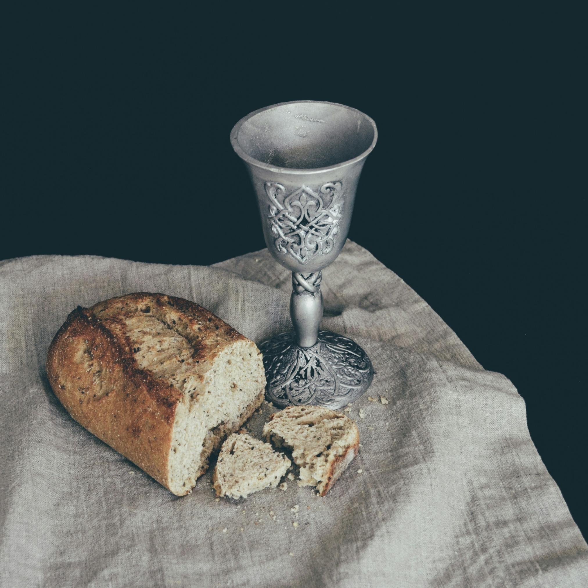 Auf einem Leinentuch liegt ein angeschnittener Laib Brot. Daneben steht ein Kelch mit Verzierungen in mattem Silber