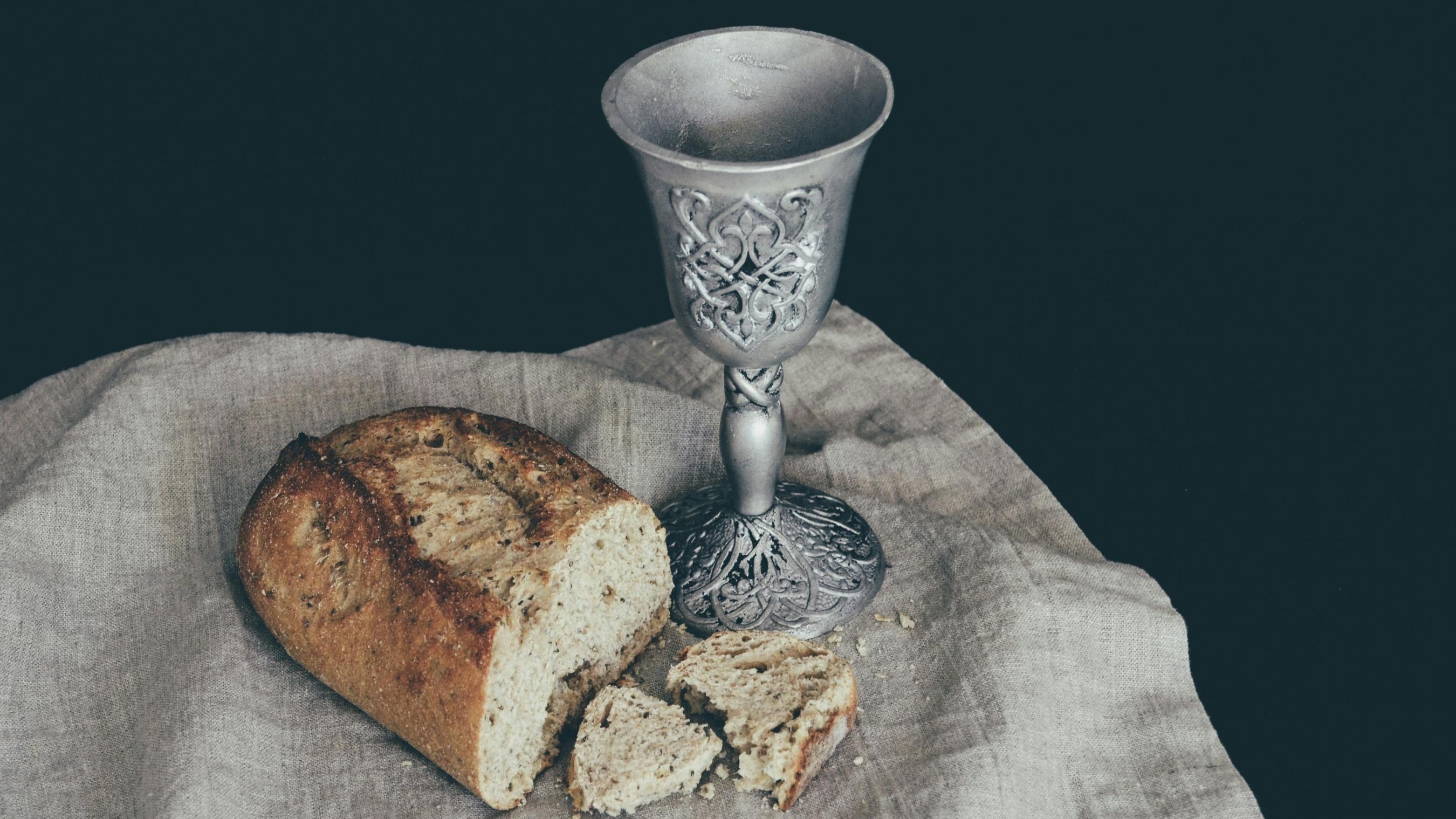 Auf einem Leinentuch liegt ein angeschnittener Laib Brot. Daneben steht ein Kelch mit Verzierungen in mattem Silber
