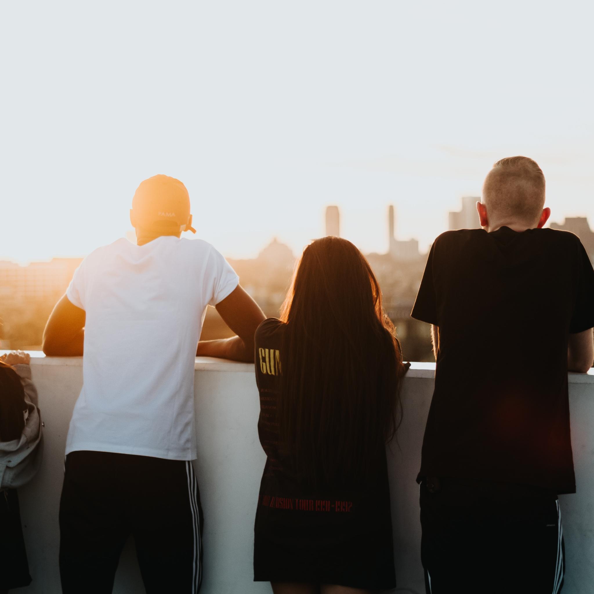 Man sieht vier Jugendliche von hinten, die sich in den Armen liegen. Sie stehen auf einem Hochhausdach und schauen in die Ferne, umgeben von warmem Licht der untergehenden Sonne.
