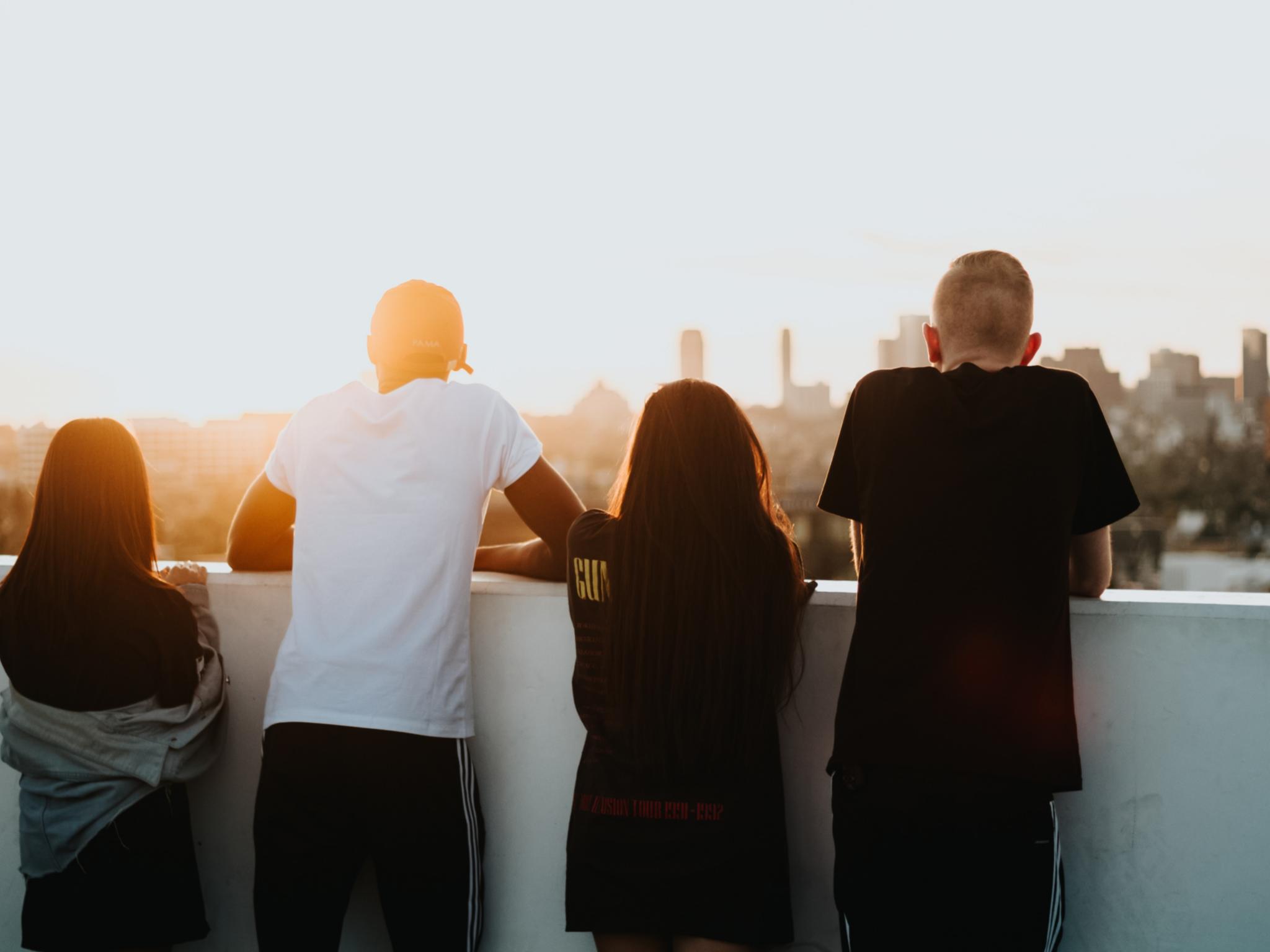 Man sieht vier Jugendliche von hinten, die sich in den Armen liegen. Sie stehen auf einem Hochhausdach und schauen in die Ferne, umgeben von warmem Licht der untergehenden Sonne.