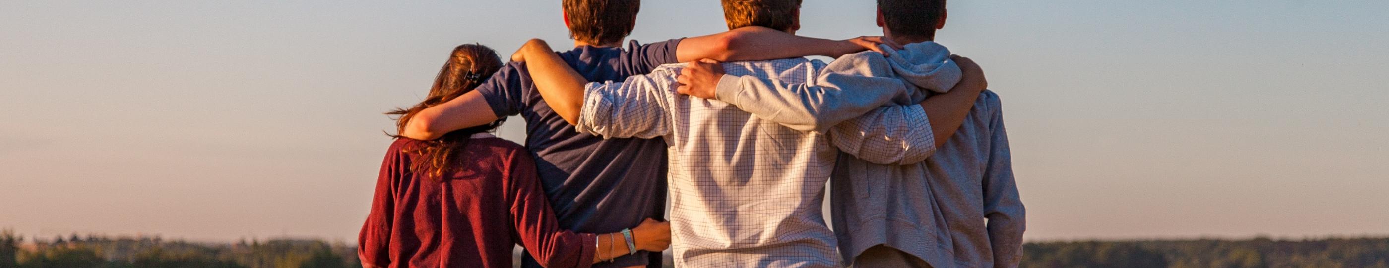 Man sieht vier Jugendliche von hinten, die auf einem Feld sitzen und sich in den Armen liegen. Es ist Sommer, der Himmel ist blau.