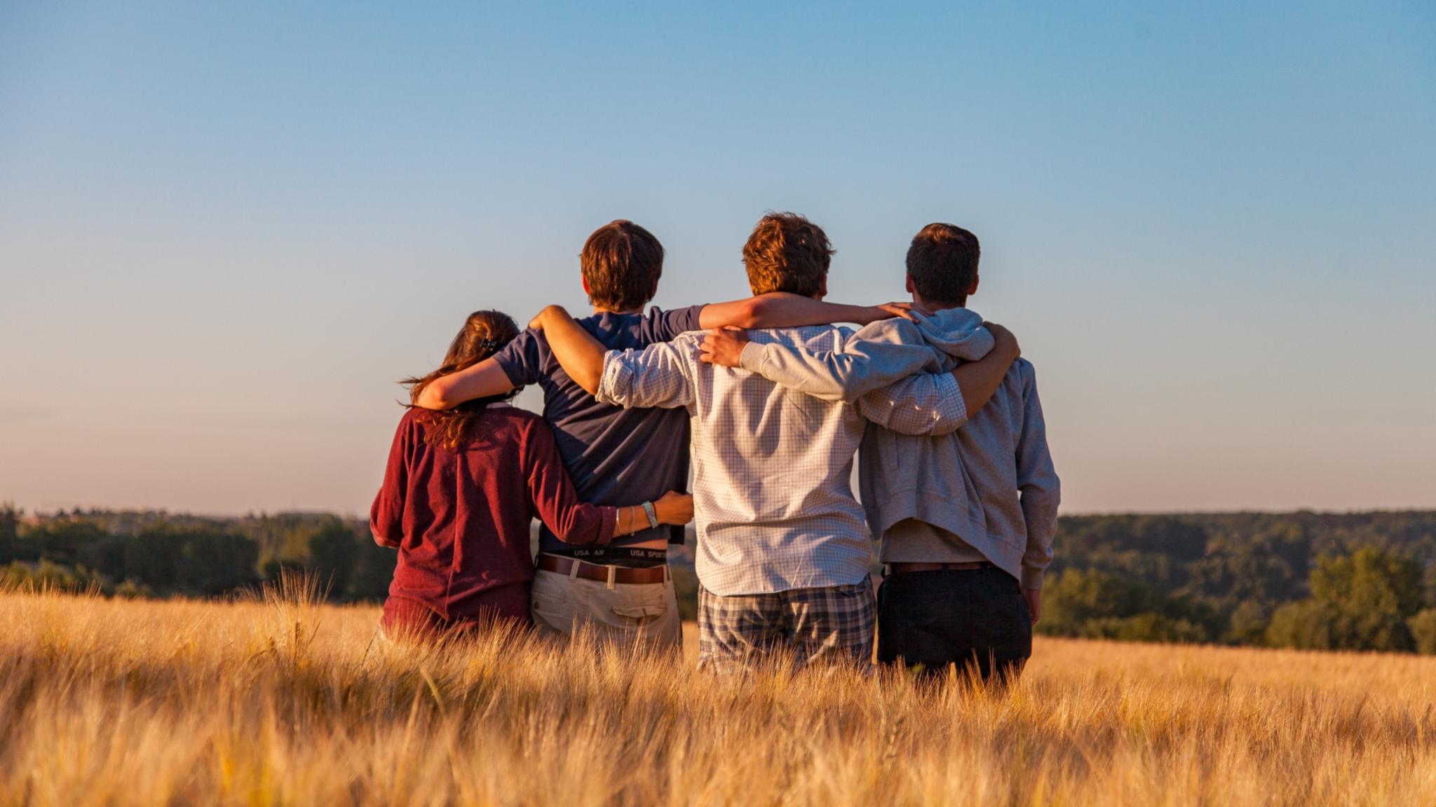 Man sieht vier Jugendliche von hinten, die auf einem Feld sitzen und sich in den Armen liegen. Es ist Sommer, der Himmel ist blau.