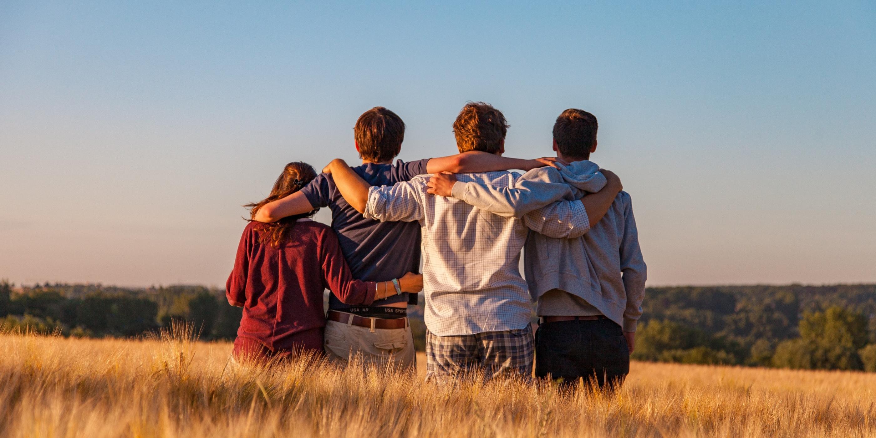 Man sieht vier Jugendliche von hinten, die auf einem Feld sitzen und sich in den Armen liegen. Es ist Sommer, der Himmel ist blau.