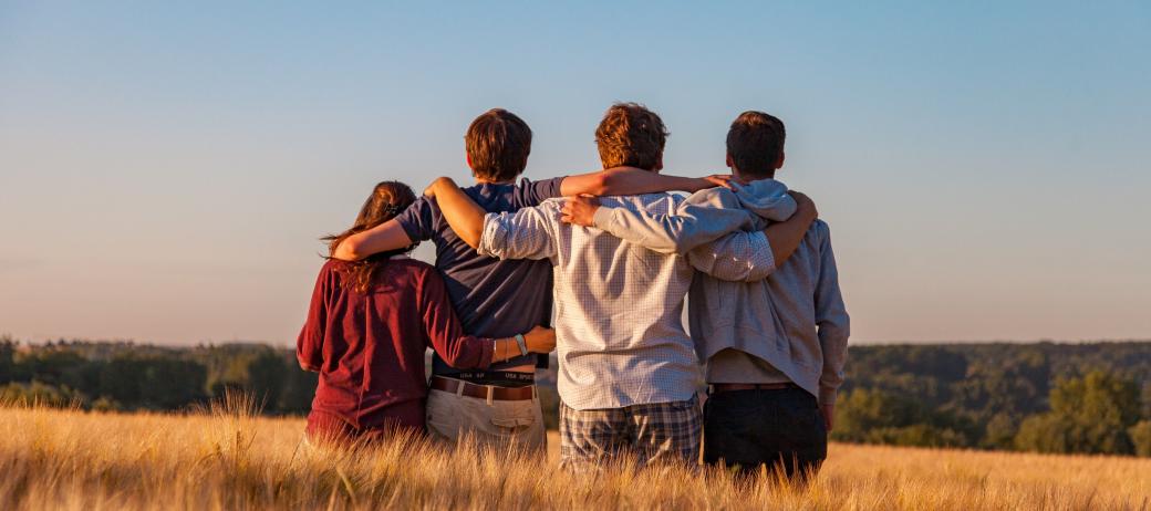 Man sieht vier Jugendliche von hinten, die auf einem Feld sitzen und sich in den Armen liegen. Es ist Sommer, der Himmel ist blau.