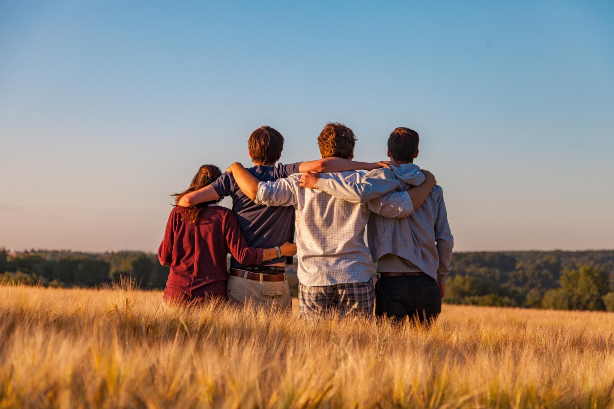 Man sieht vier Jugendliche von hinten, die auf einem Feld sitzen und sich in den Armen liegen. Es ist Sommer, der Himmel ist blau.