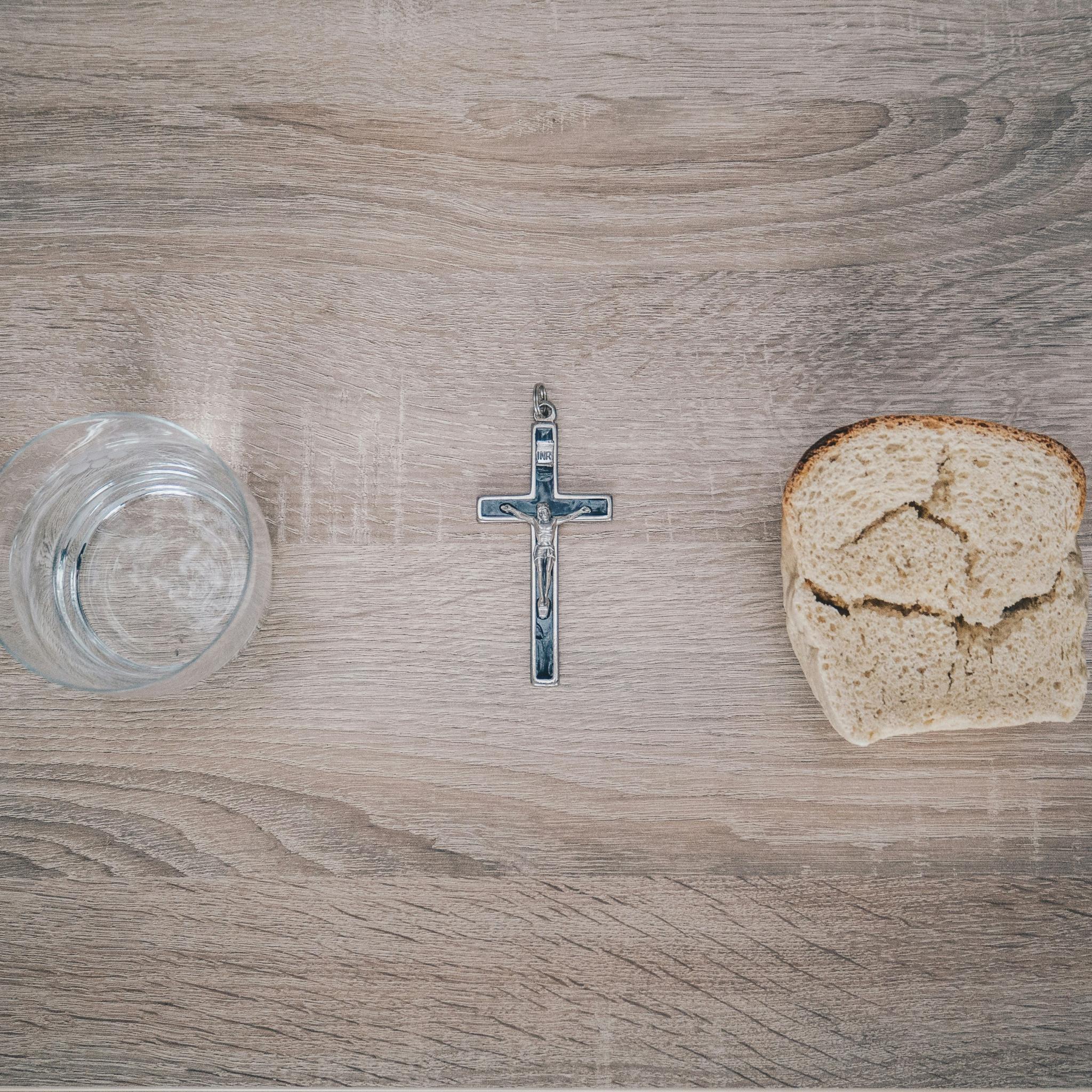 Blick von oben auf einen Tisch. Links steht ein Glas mit Wasser, rechts liegt eine Scheibe Brot. Dazwischen liegt ein Kreuz