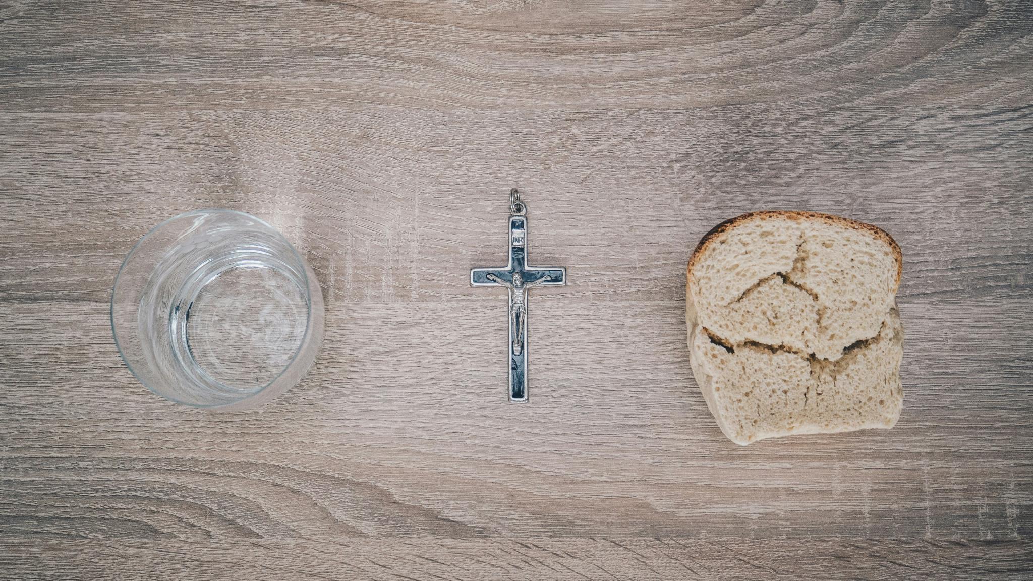 Blick von oben auf einen Tisch. Links steht ein Glas mit Wasser, rechts liegt eine Scheibe Brot. Dazwischen liegt ein Kreuz