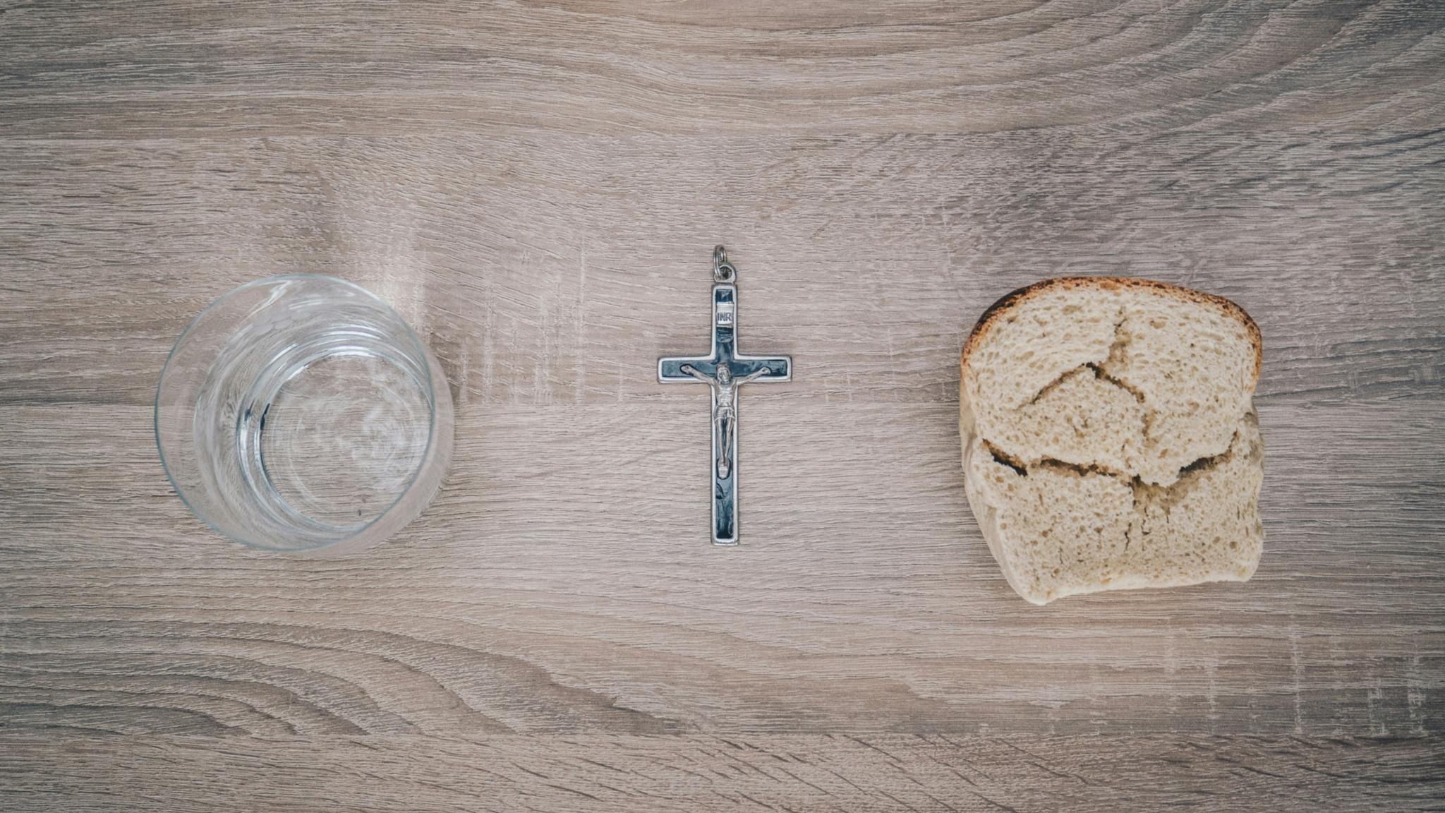 Blick von oben auf einen Tisch. Links steht ein Glas mit Wasser, rechts liegt eine Scheibe Brot. Dazwischen liegt ein Kreuz