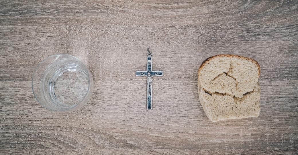 Blick von oben auf einen Tisch. Links steht ein Glas mit Wasser, rechts liegt eine Scheibe Brot. Dazwischen liegt ein Kreuz