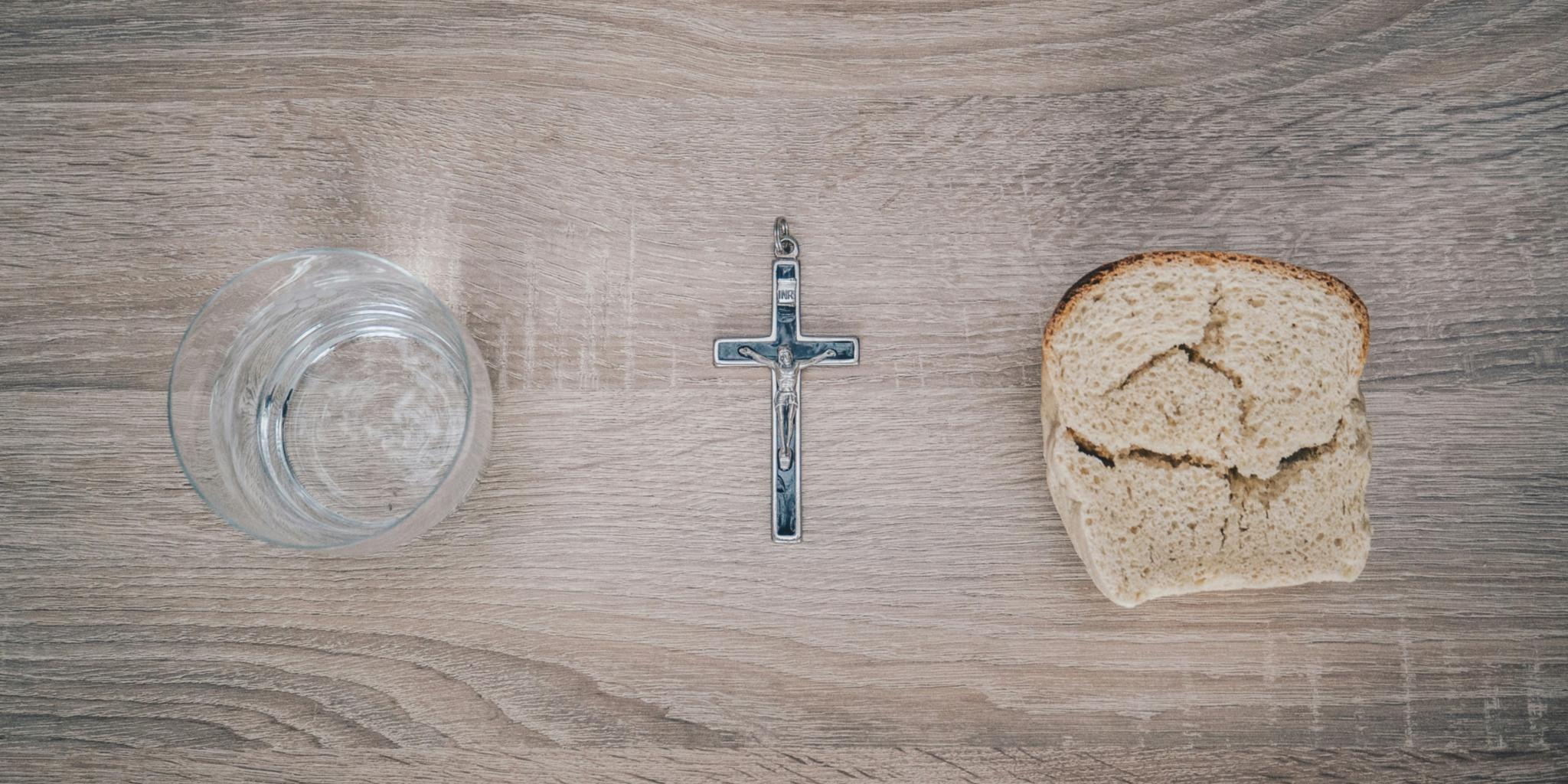 Blick von oben auf einen Tisch. Links steht ein Glas mit Wasser, rechts liegt eine Scheibe Brot. Dazwischen liegt ein Kreuz