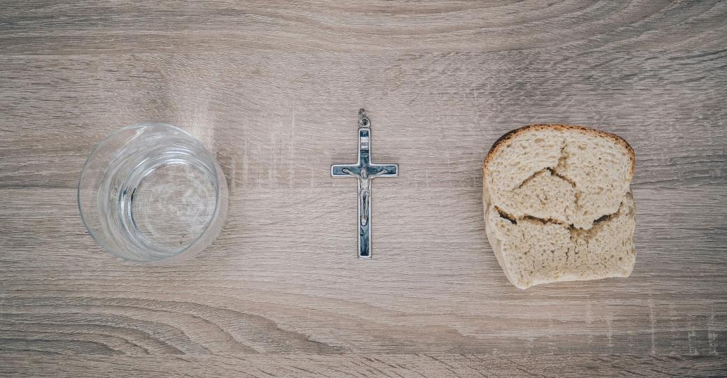 Blick von oben auf einen Tisch. Links steht ein Glas mit Wasser, rechts liegt eine Scheibe Brot. Dazwischen liegt ein Kreuz