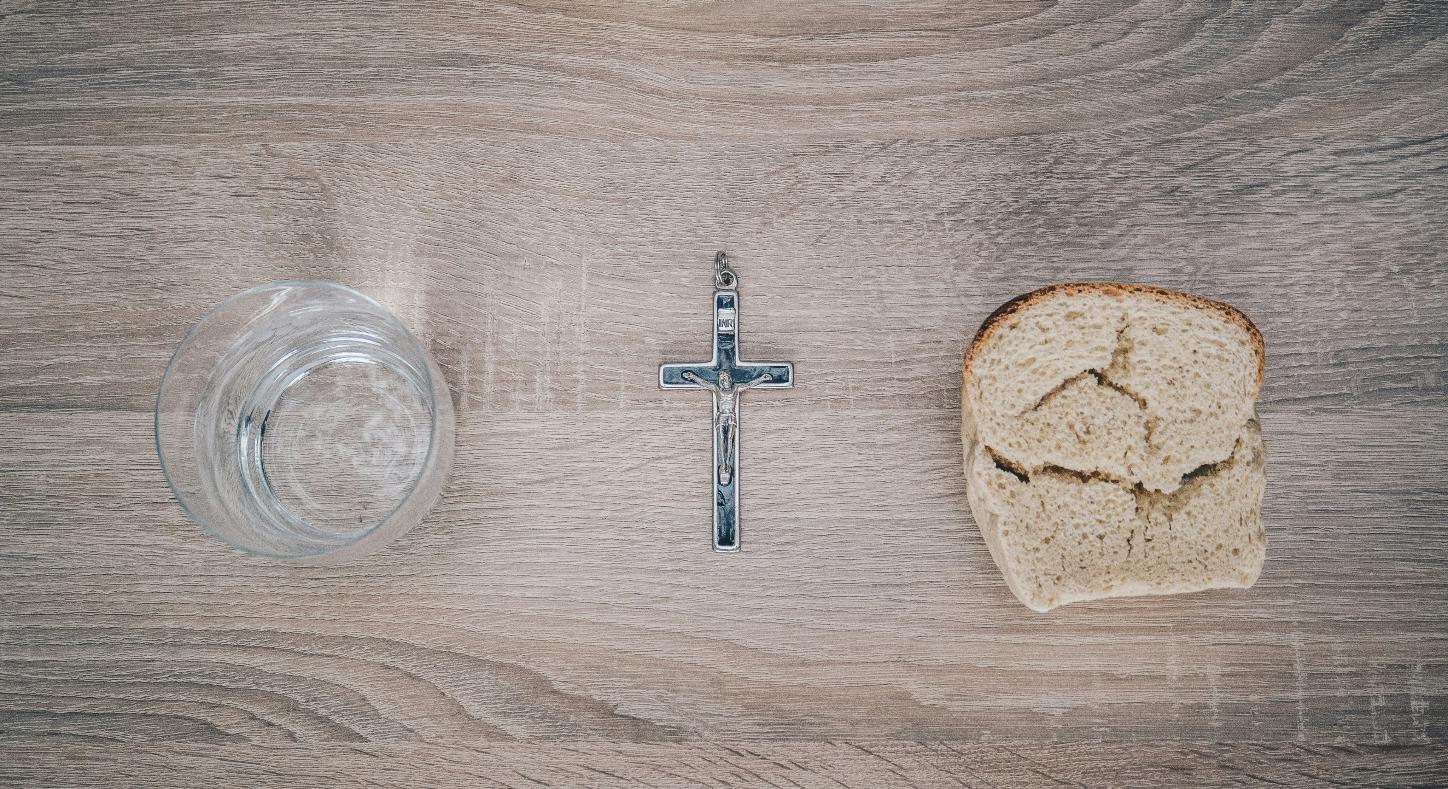 Blick von oben auf einen Tisch. Links steht ein Glas mit Wasser, rechts liegt eine Scheibe Brot. Dazwischen liegt ein Kreuz