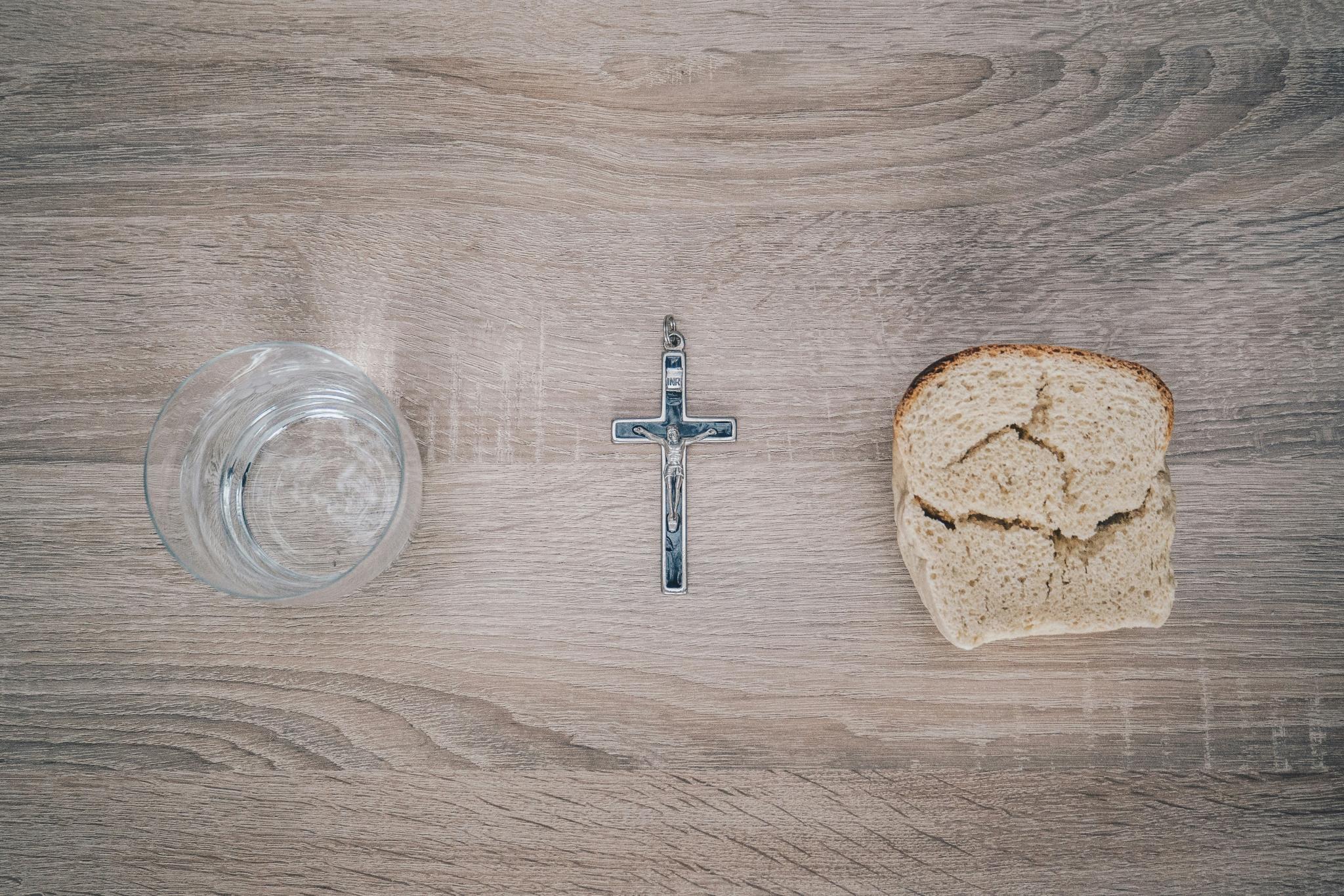 Blick von oben auf einen Tisch. Links steht ein Glas mit Wasser, rechts liegt eine Scheibe Brot. Dazwischen liegt ein Kreuz