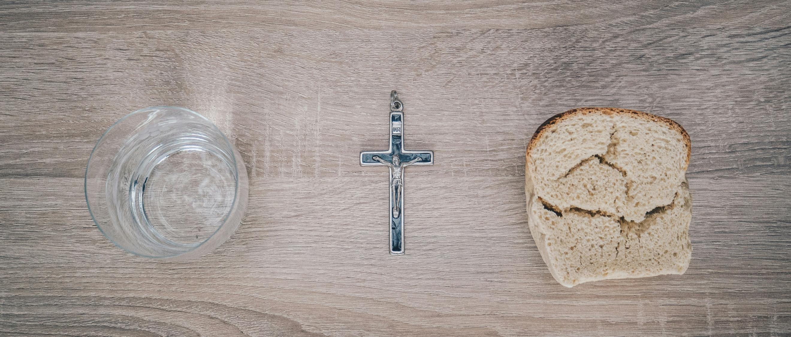 Blick von oben auf einen Tisch. Links steht ein Glas mit Wasser, rechts liegt eine Scheibe Brot. Dazwischen liegt ein Kreuz