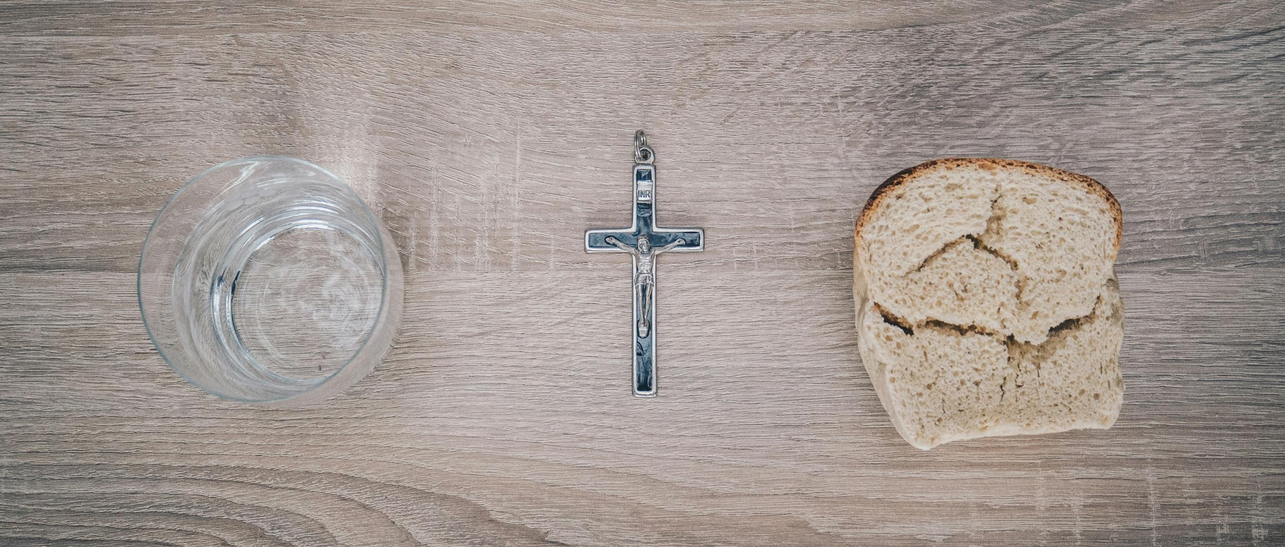 Blick von oben auf einen Tisch. Links steht ein Glas mit Wasser, rechts liegt eine Scheibe Brot. Dazwischen liegt ein Kreuz