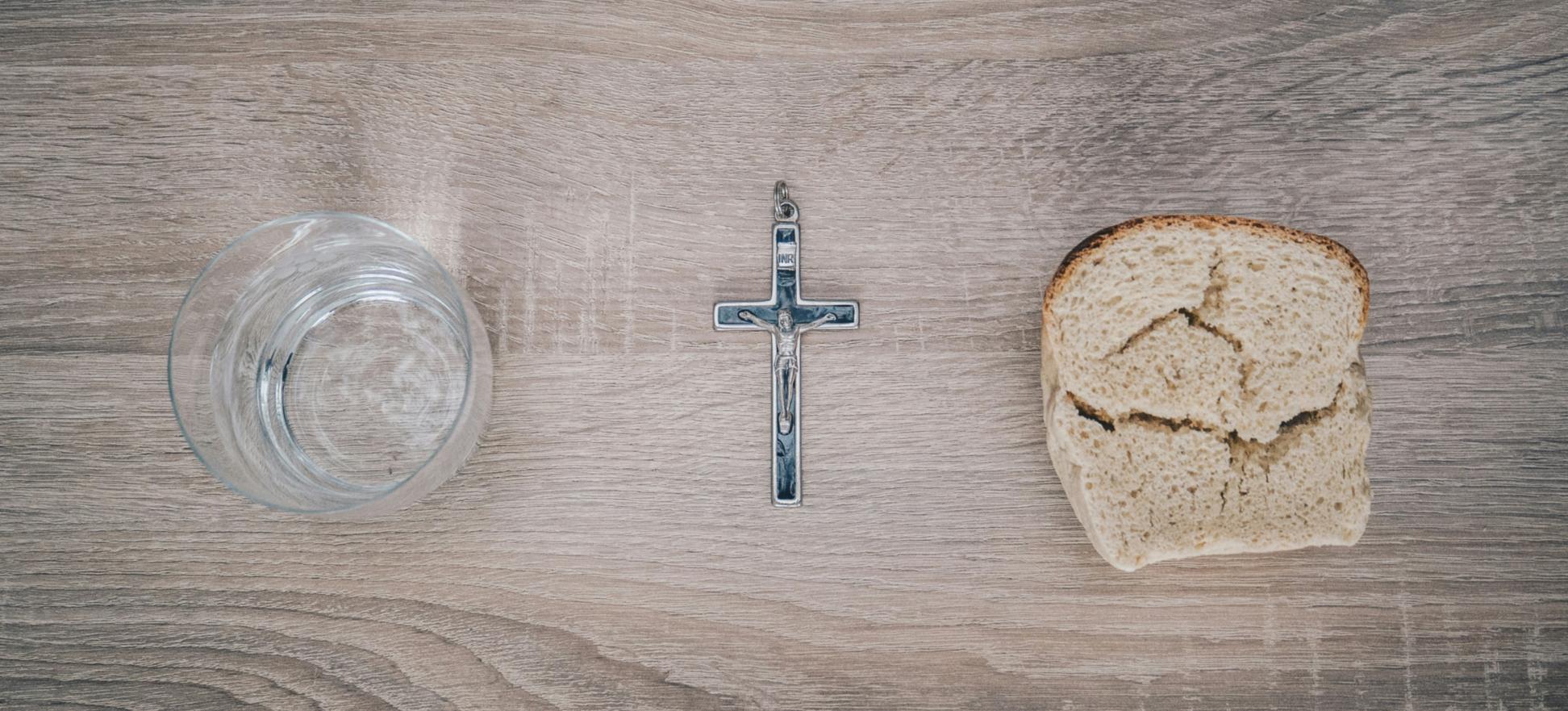 Blick von oben auf einen Tisch. Links steht ein Glas mit Wasser, rechts liegt eine Scheibe Brot. Dazwischen liegt ein Kreuz