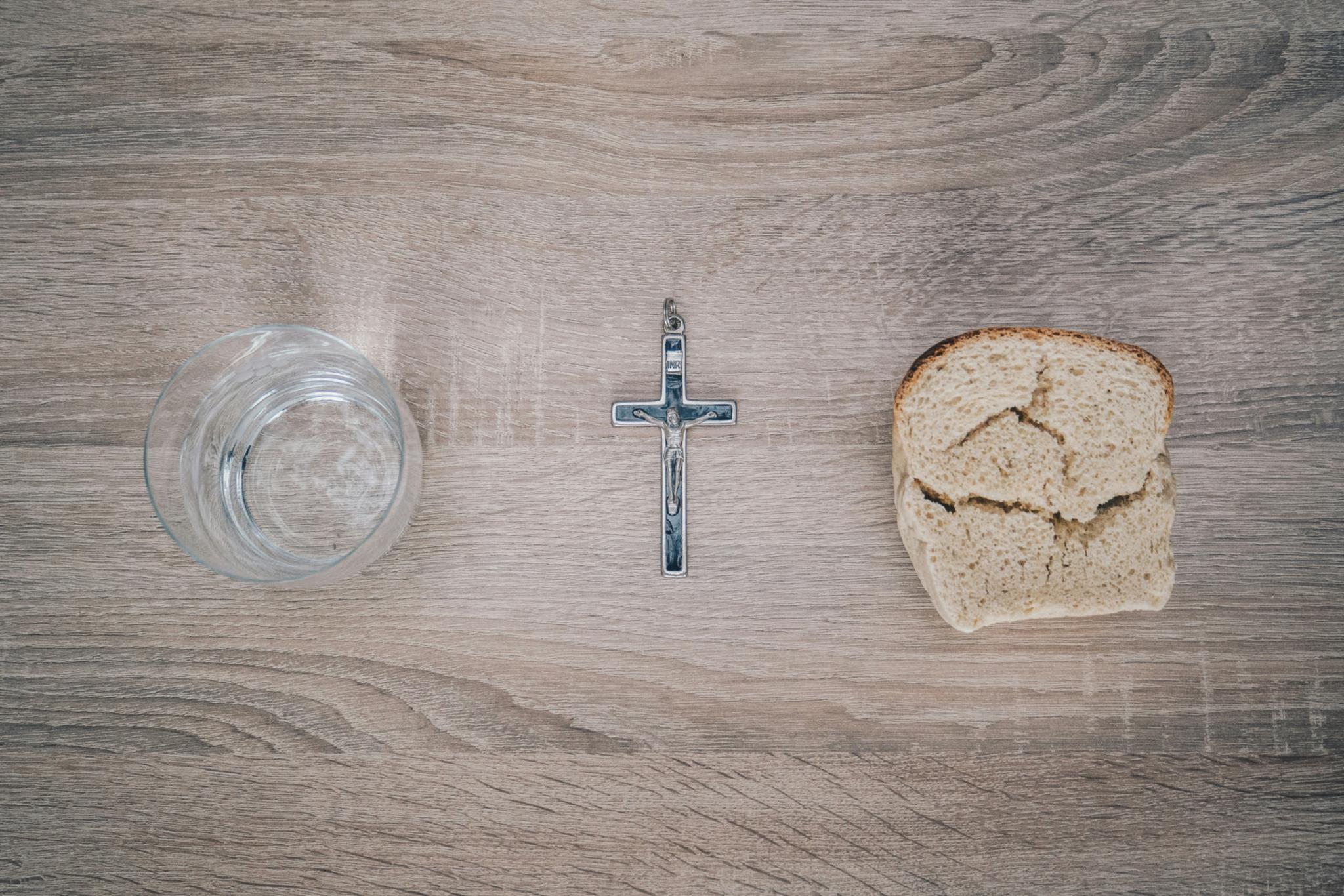 Blick von oben auf einen Tisch. Links steht ein Glas mit Wasser, rechts liegt eine Scheibe Brot. Dazwischen liegt ein Kreuz