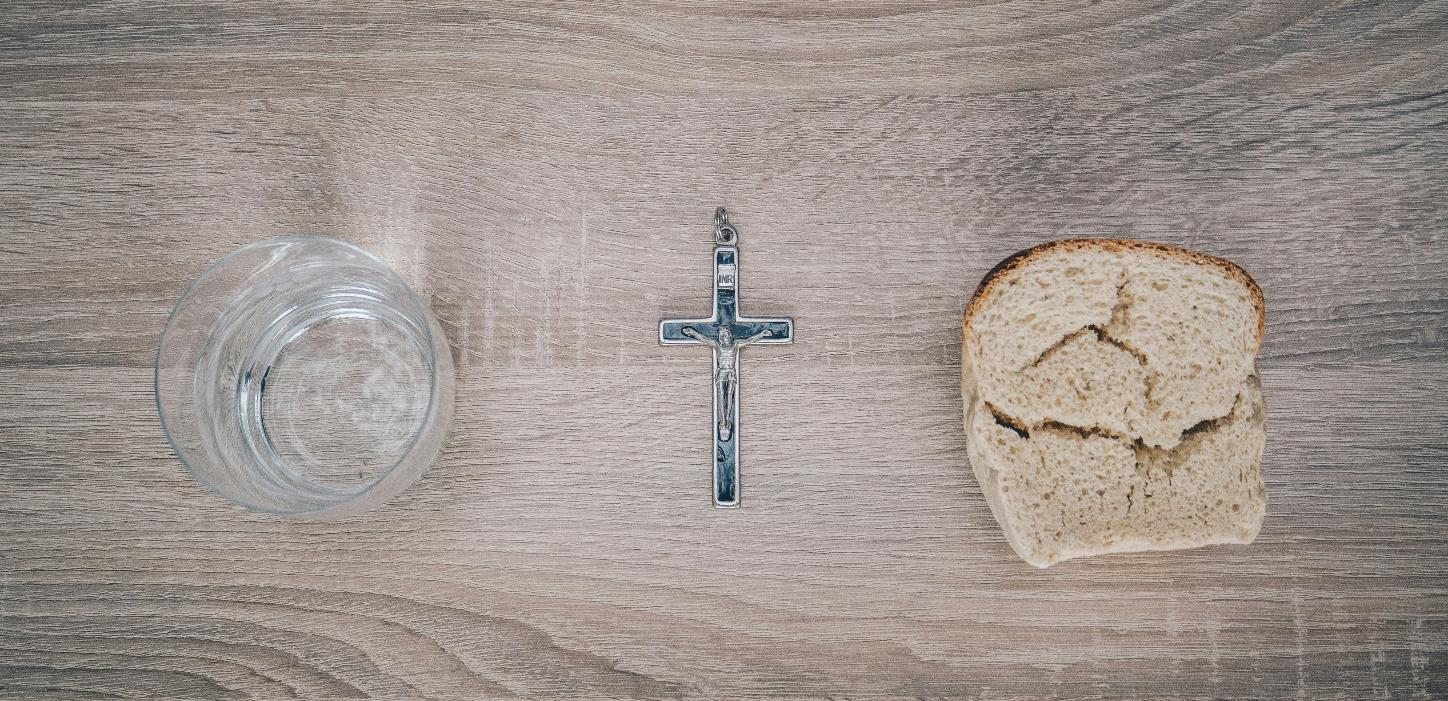 Blick von oben auf einen Tisch. Links steht ein Glas mit Wasser, rechts liegt eine Scheibe Brot. Dazwischen liegt ein Kreuz