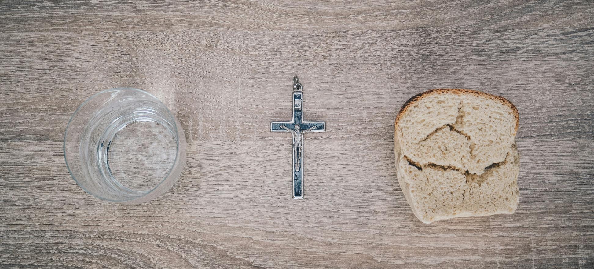 Blick von oben auf einen Tisch. Links steht ein Glas mit Wasser, rechts liegt eine Scheibe Brot. Dazwischen liegt ein Kreuz