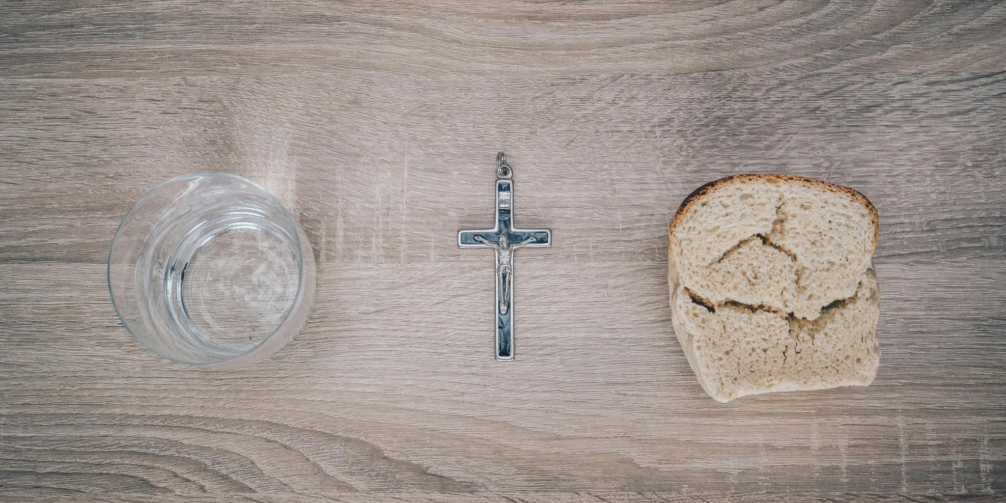 Blick von oben auf einen Tisch. Links steht ein Glas mit Wasser, rechts liegt eine Scheibe Brot. Dazwischen liegt ein Kreuz