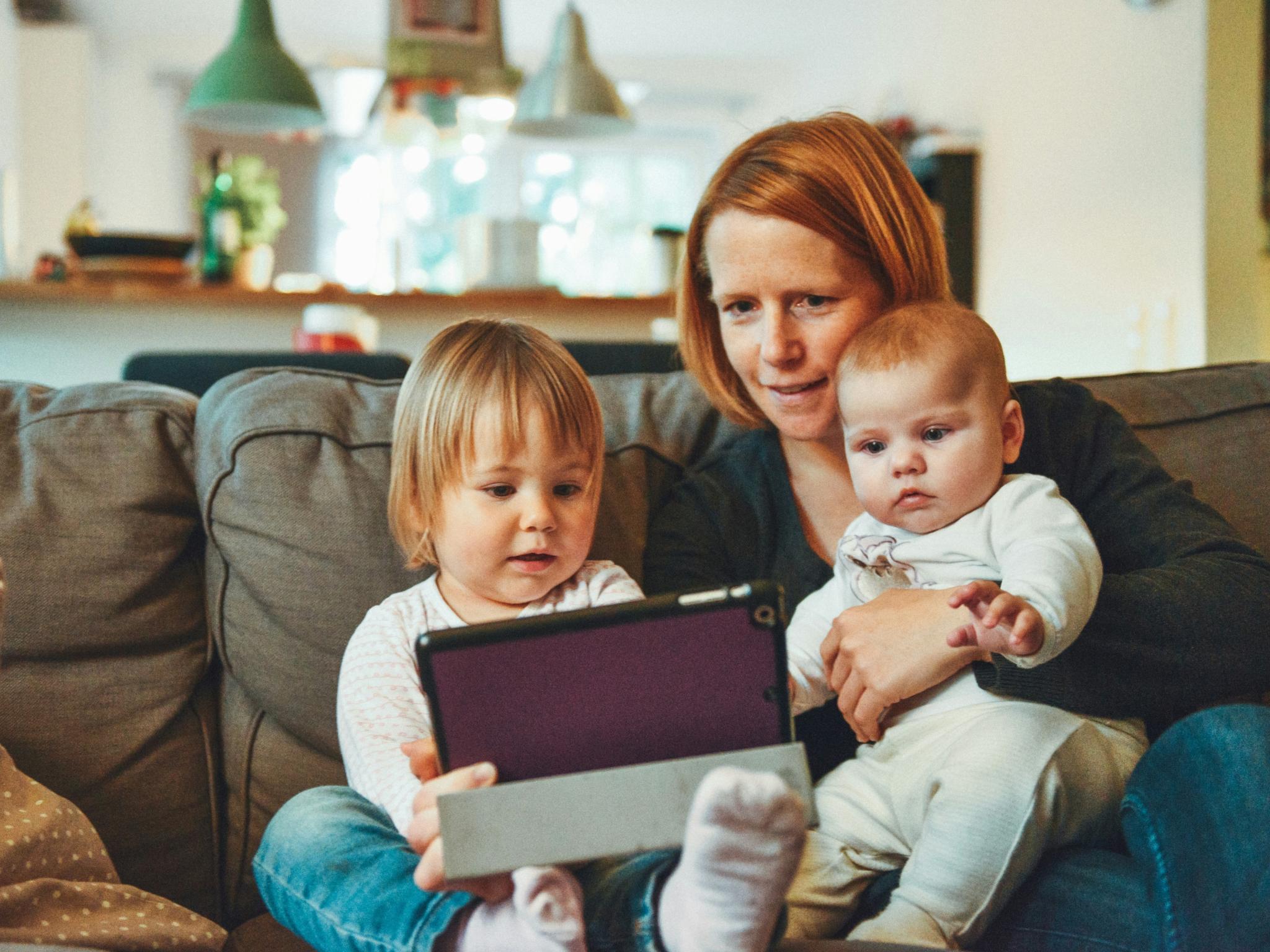 Eine Frau sitzt mit Baby und Kleinkind auf einem Sofa. Alle drei schauen auf ein Tablet