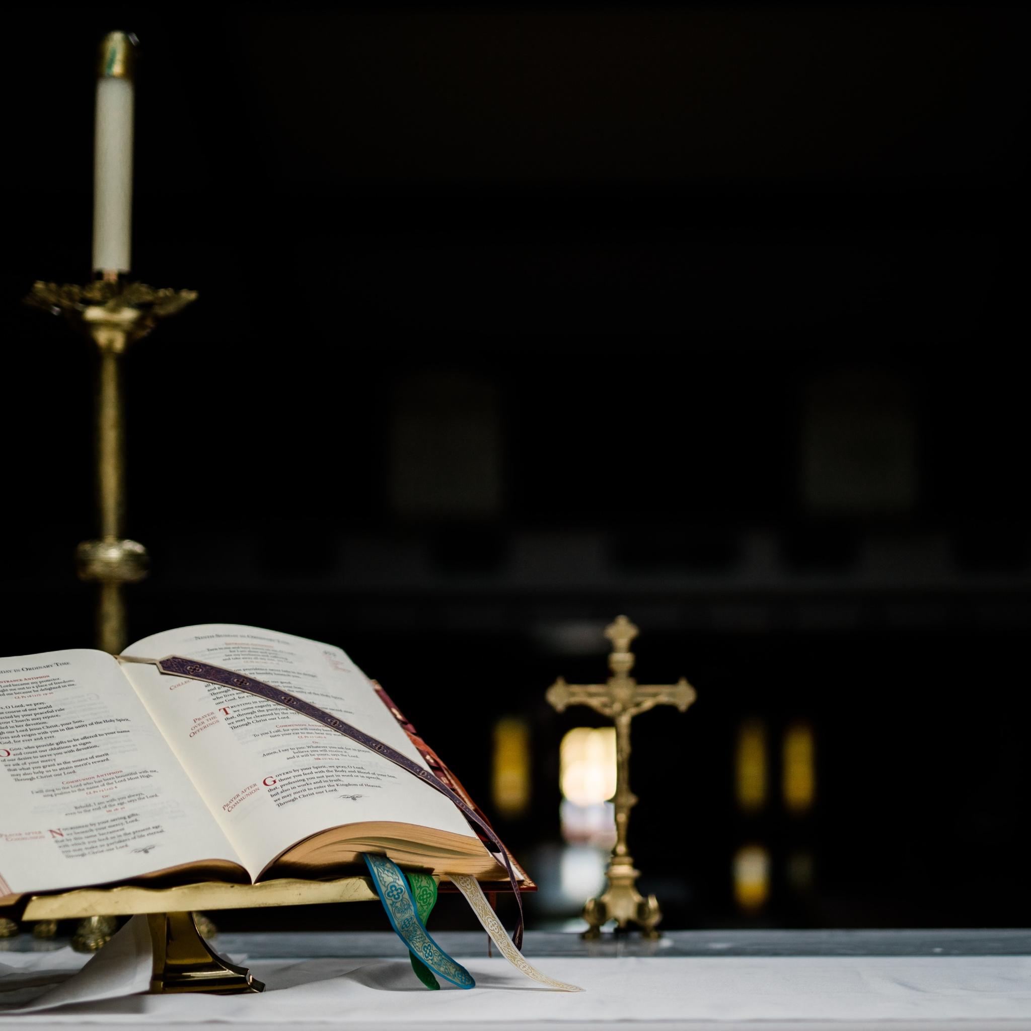 Auf einem Altar steht ein geöffnetes Buch in einem Halter, dahinter ein kleines Kreuz und mehrere große Kerzenständer