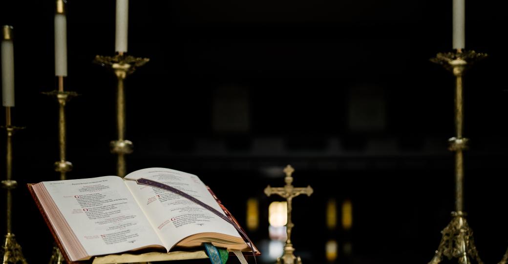 Auf einem Altar steht ein geöffnetes Buch in einem Halter, dahinter ein kleines Kreuz und mehrere große Kerzenständer