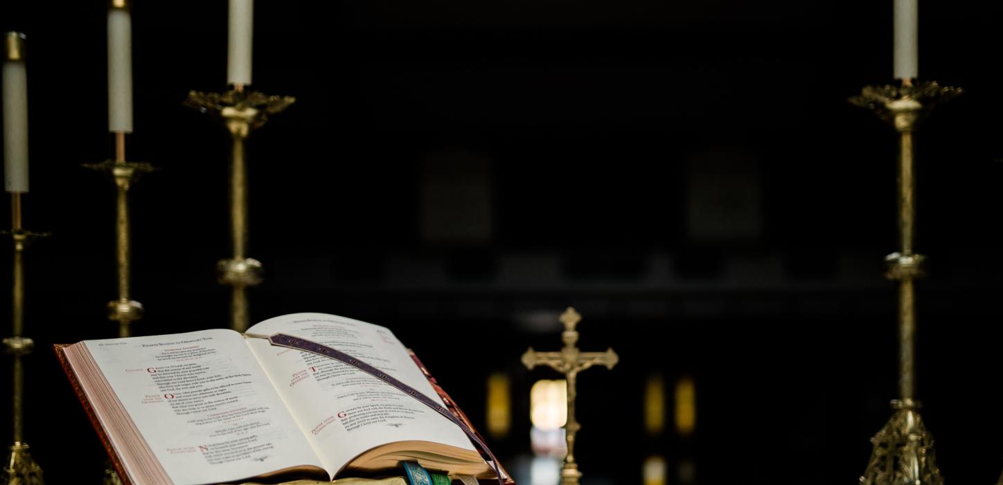 Auf einem Altar steht ein geöffnetes Buch in einem Halter, dahinter ein kleines Kreuz und mehrere große Kerzenständer