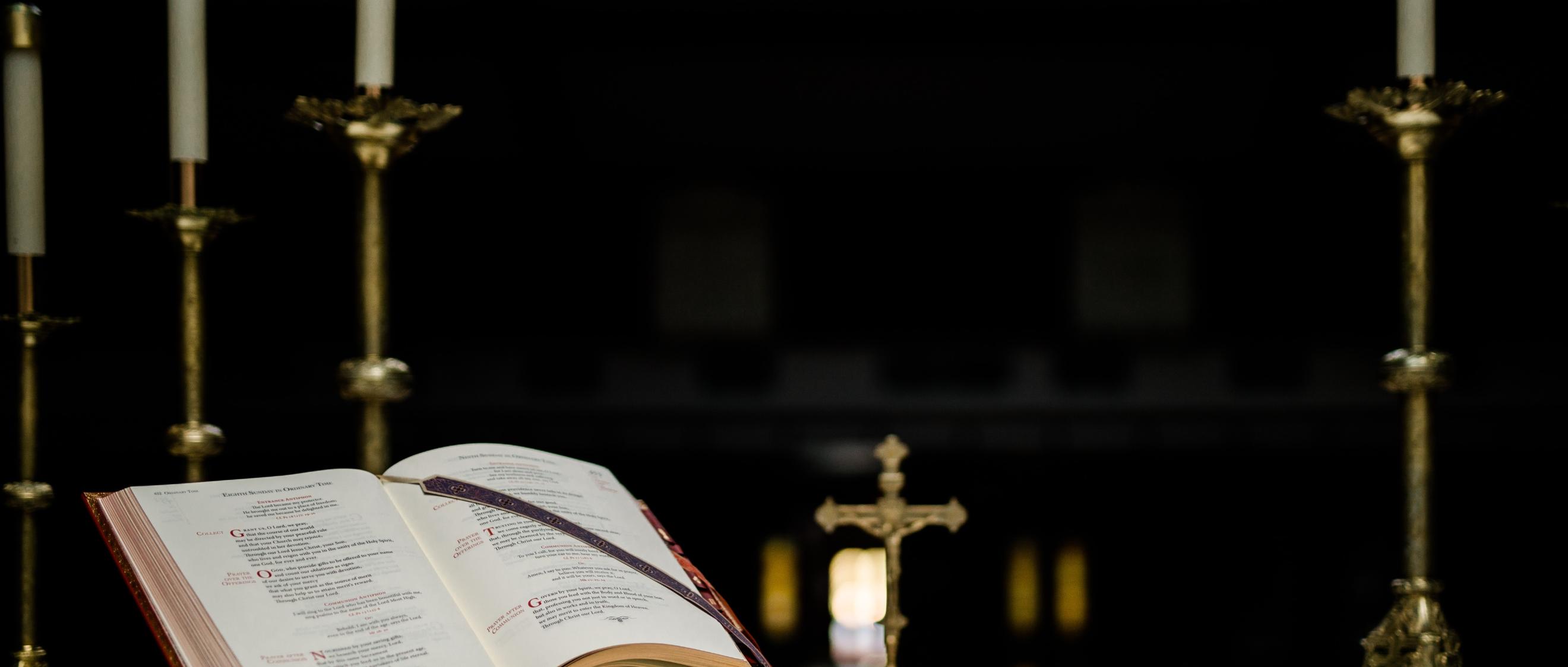 Auf einem Altar steht ein geöffnetes Buch in einem Halter, dahinter ein kleines Kreuz und mehrere große Kerzenständer