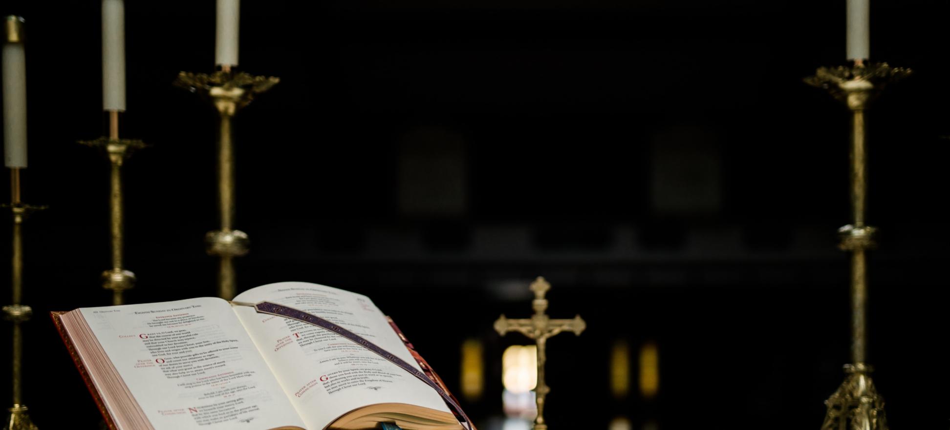 Auf einem Altar steht ein geöffnetes Buch in einem Halter, dahinter ein kleines Kreuz und mehrere große Kerzenständer