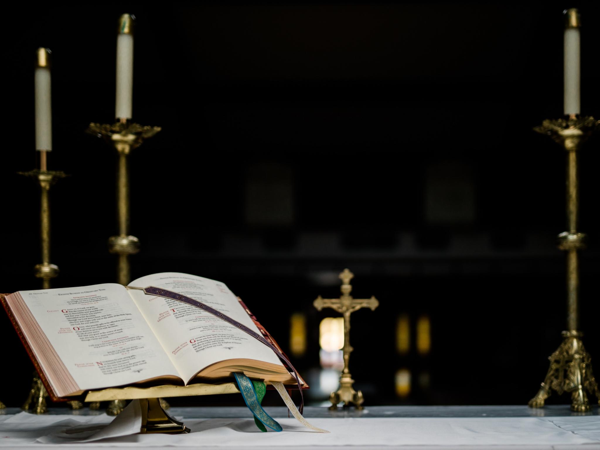 Auf einem Altar steht ein geöffnetes Buch in einem Halter, dahinter ein kleines Kreuz und mehrere große Kerzenständer