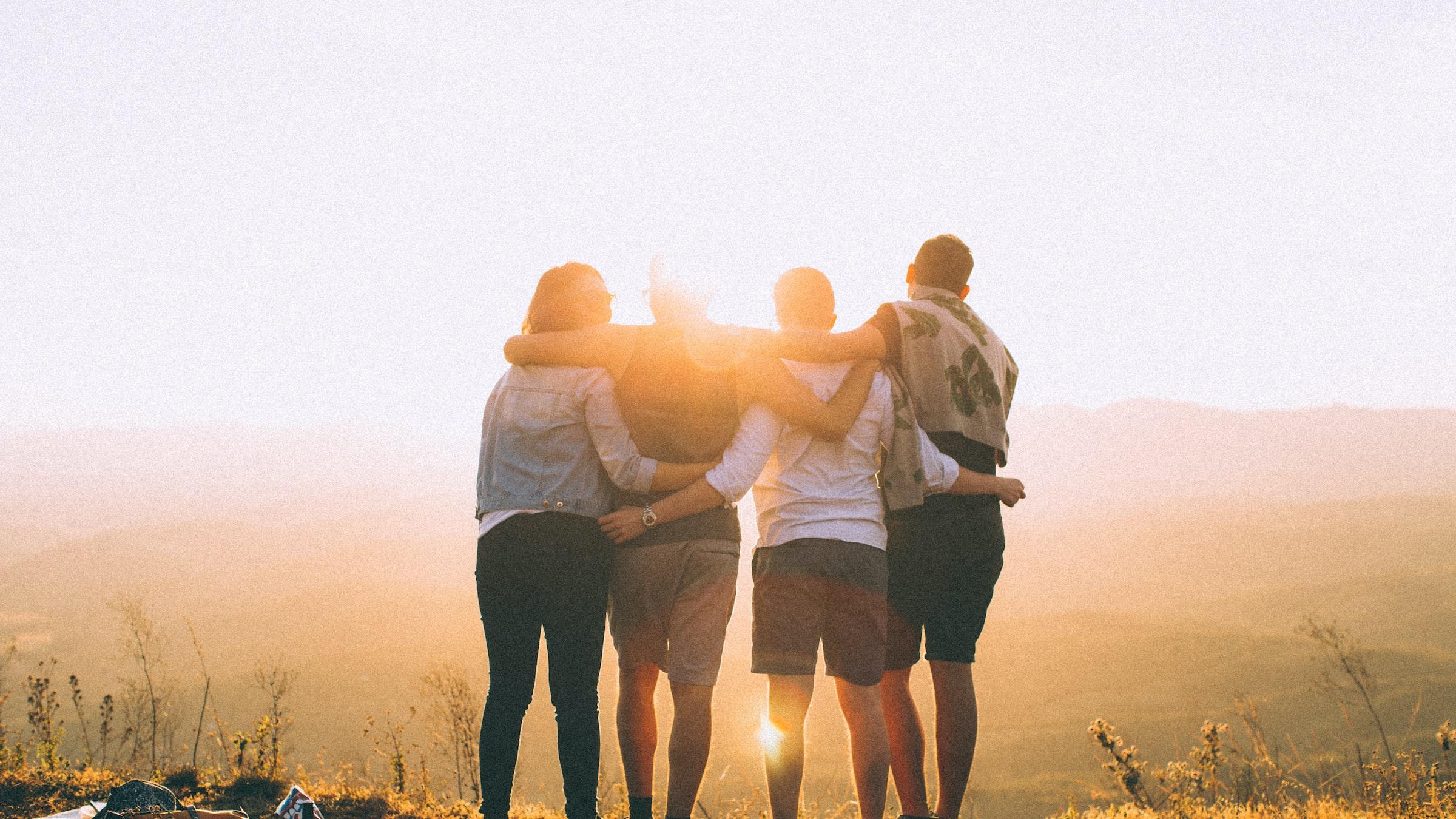 Vier Jugendliche stehen auf einem Hügel, halten sich in den Armen und schauen in die Ferne - umgeben vom warmen Licht der untergehenden Sonne.