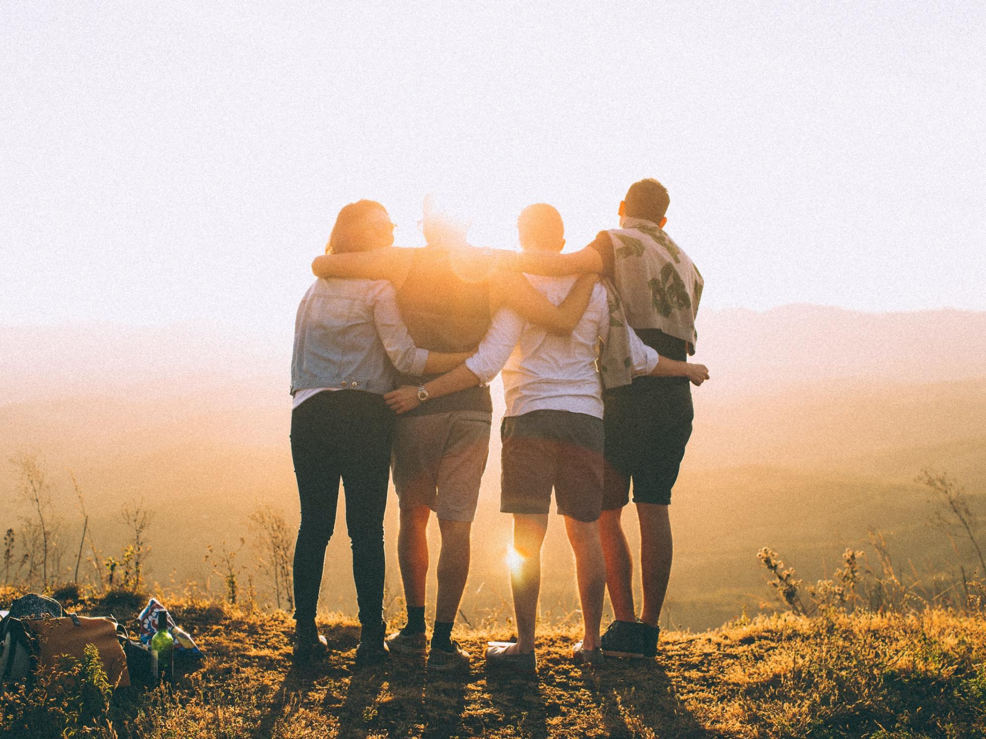 Vier Jugendliche stehen auf einem Hügel, halten sich in den Armen und schauen in die Ferne - umgeben vom warmen Licht der untergehenden Sonne.