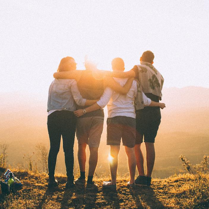 Vier Jugendliche stehen auf einem Hügel, halten sich in den Armen und schauen in die Ferne - umgeben vom warmen Licht der untergehenden Sonne.