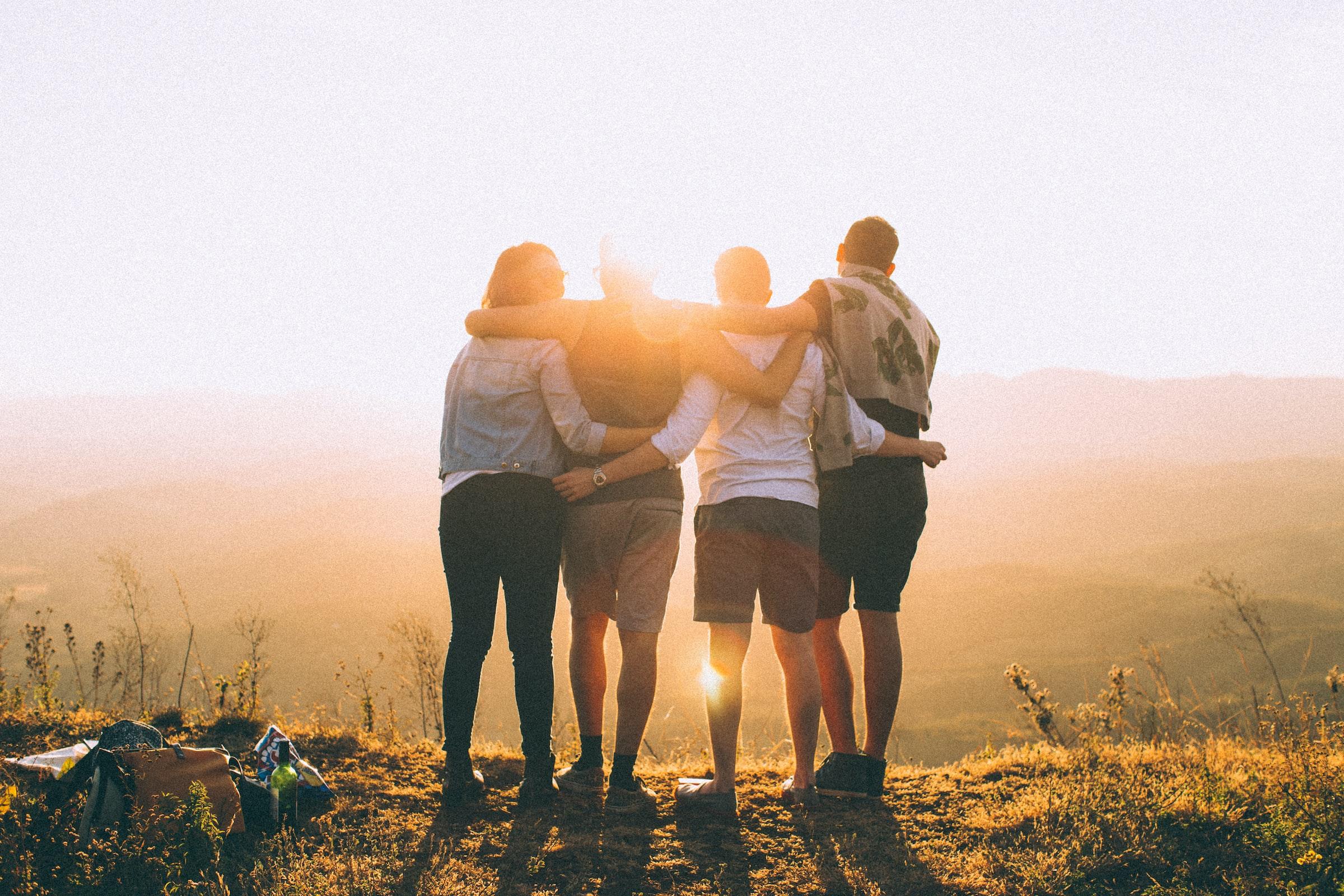 Vier Jugendliche stehen auf einem Hügel, halten sich in den Armen und schauen in die Ferne - umgeben vom warmen Licht der untergehenden Sonne.