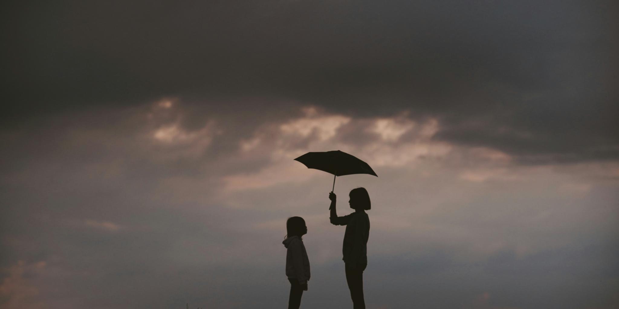 Silhouette: Erwachsene Person hält vor einem Gewitterhimmel einen Schirm über sich und ein Kind