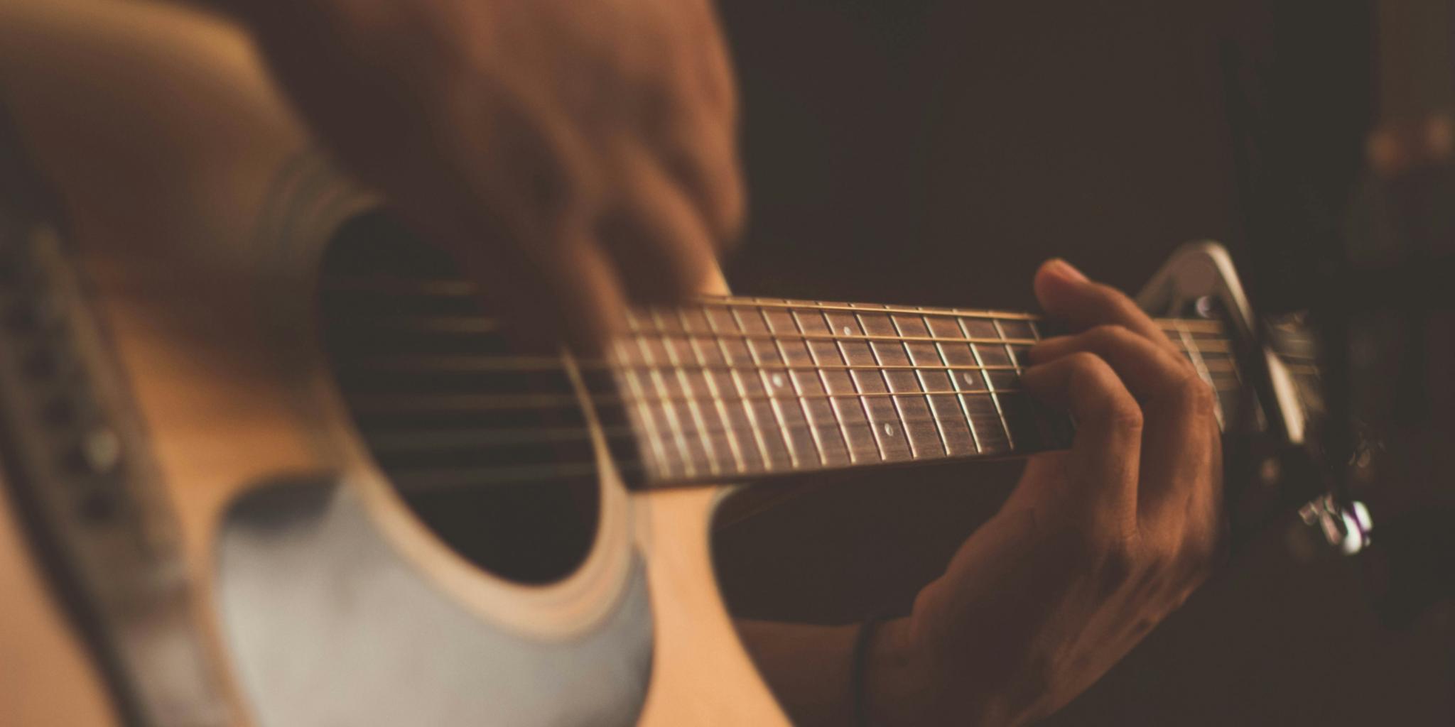 Auf dem Bild ist der Körper einer Gitarre abgebildet, die gerade von jemand gespielt wird. Eine Hand greift Akkorde am Steg und eine Hand streicht über die Seiten.