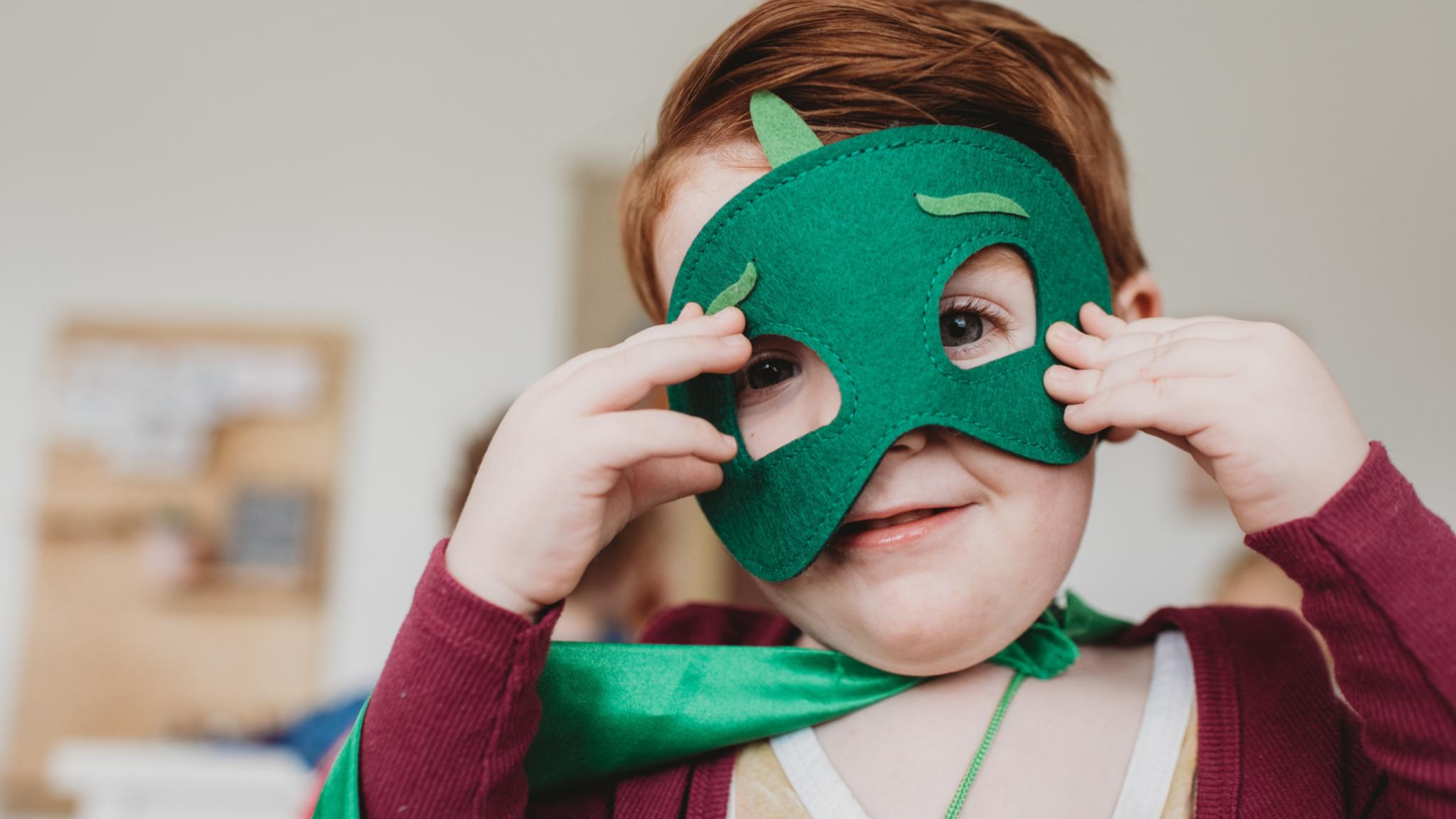 lachender kleiner junge mit grüner Superheldenmaske und grünem Umhang