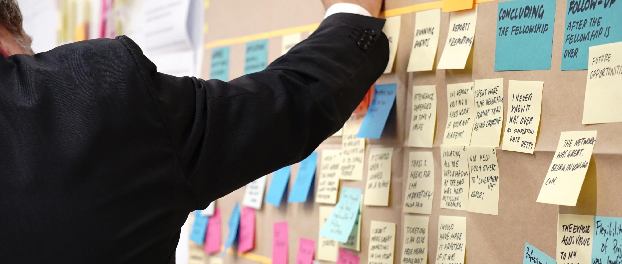Arm eines Mannes zeigt auf eine Pinnwand, an der verschiedenenfarbige Klebezettel angebracht sind.